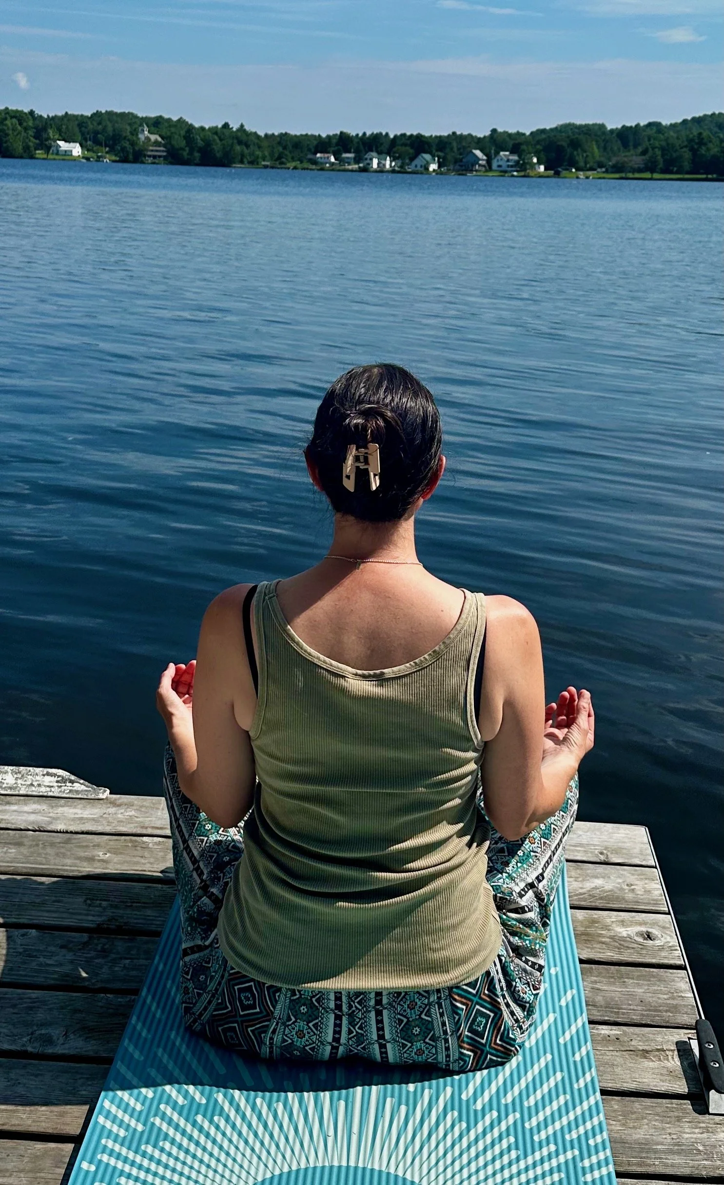 Person practicing yoga on a dock by a lake with houses and trees in the background.