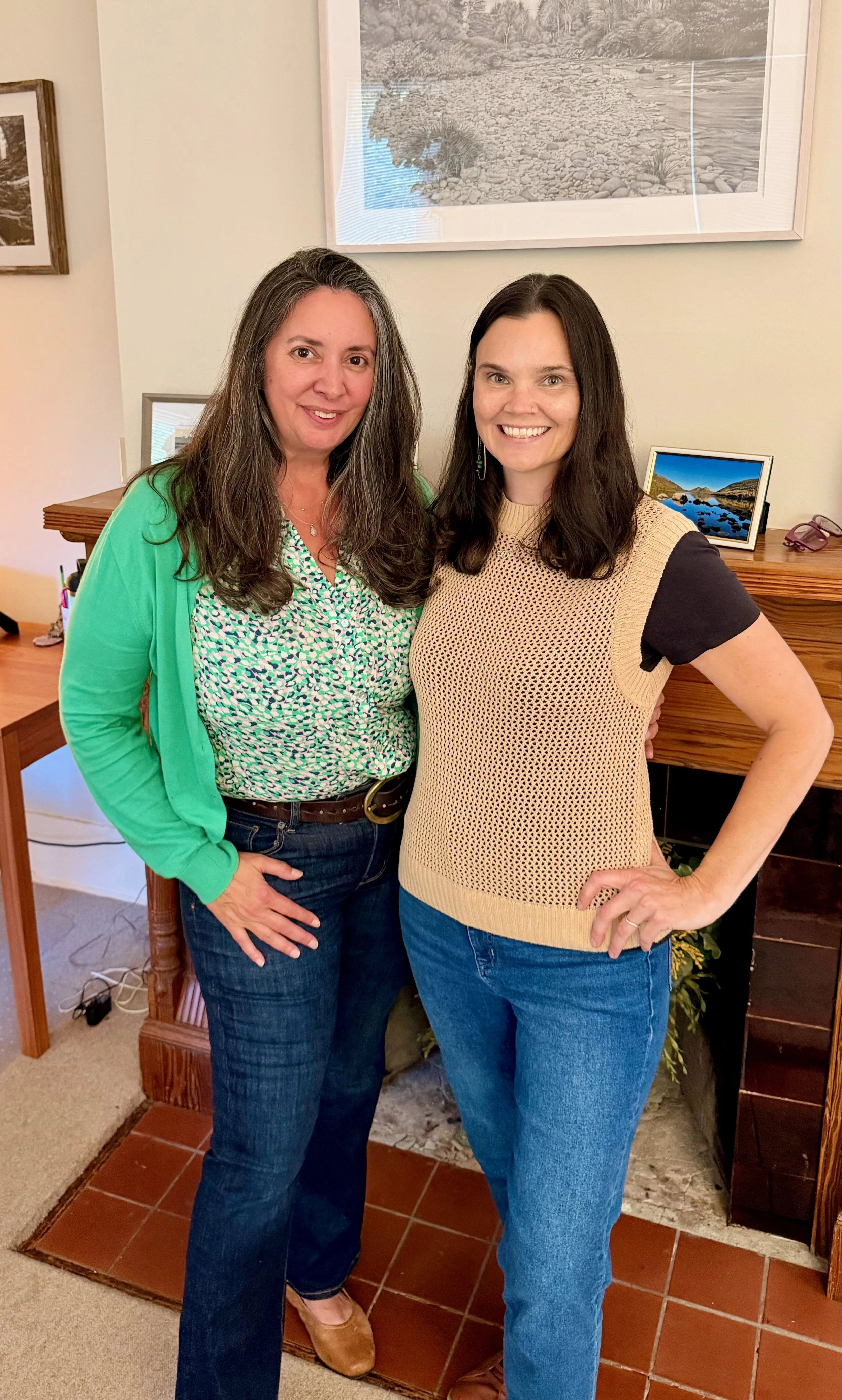 Two women standing close together indoors, smiling at the camera. One has long gray hair and is wearing a green cardigan and patterned blouse. The other has long dark hair and is wearing a beige sleeveless sweater over a black shirt. The background has framed photos and a small digital picture frame on a wooden table, with a drawing of a river landscape on the wall.
