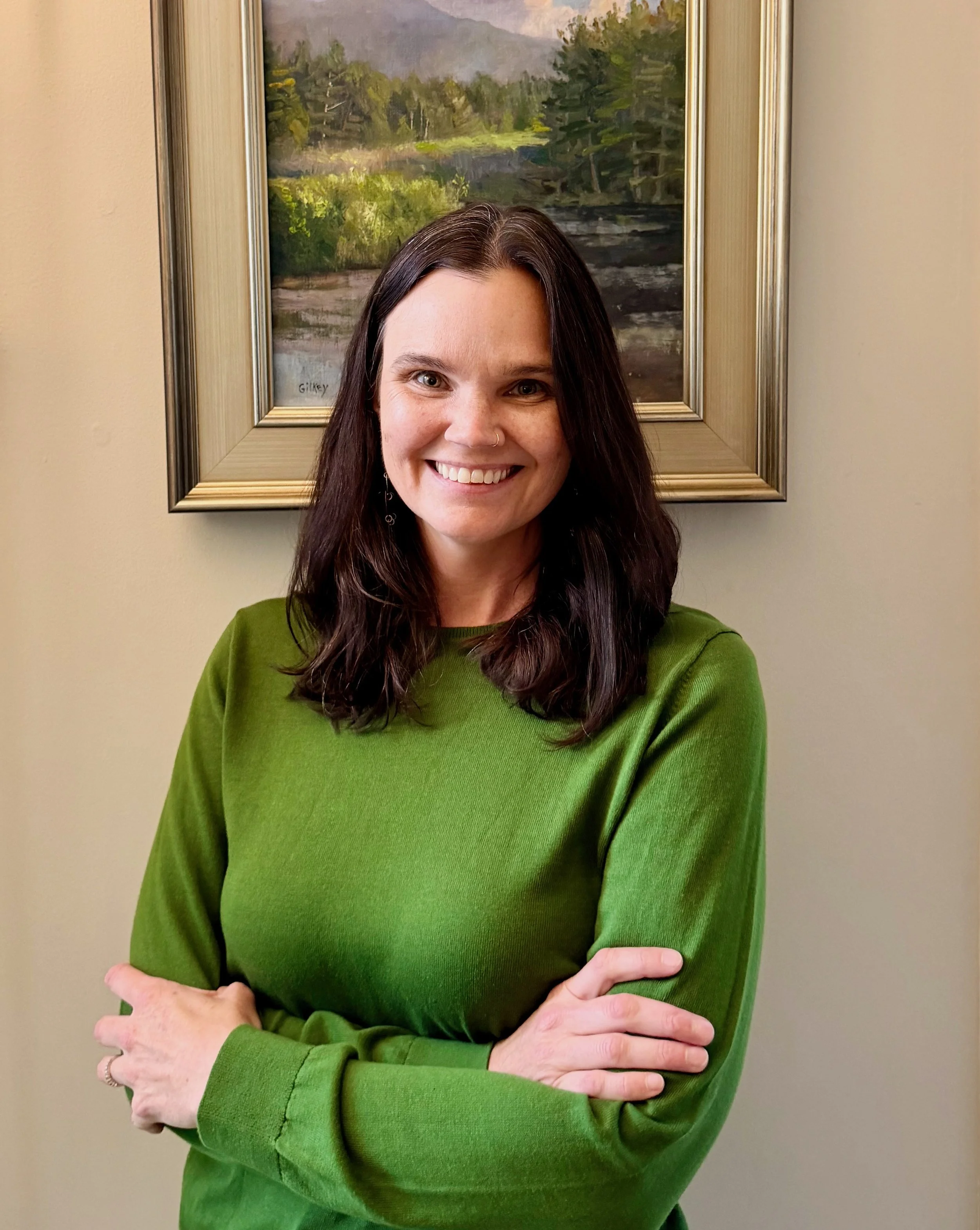 A woman with long dark hair, part of her face smiling, wearing a green sweater with her arms crossed, standing in front of a framed landscape painting of a river, trees, and a cloudy sky on a beige wall.