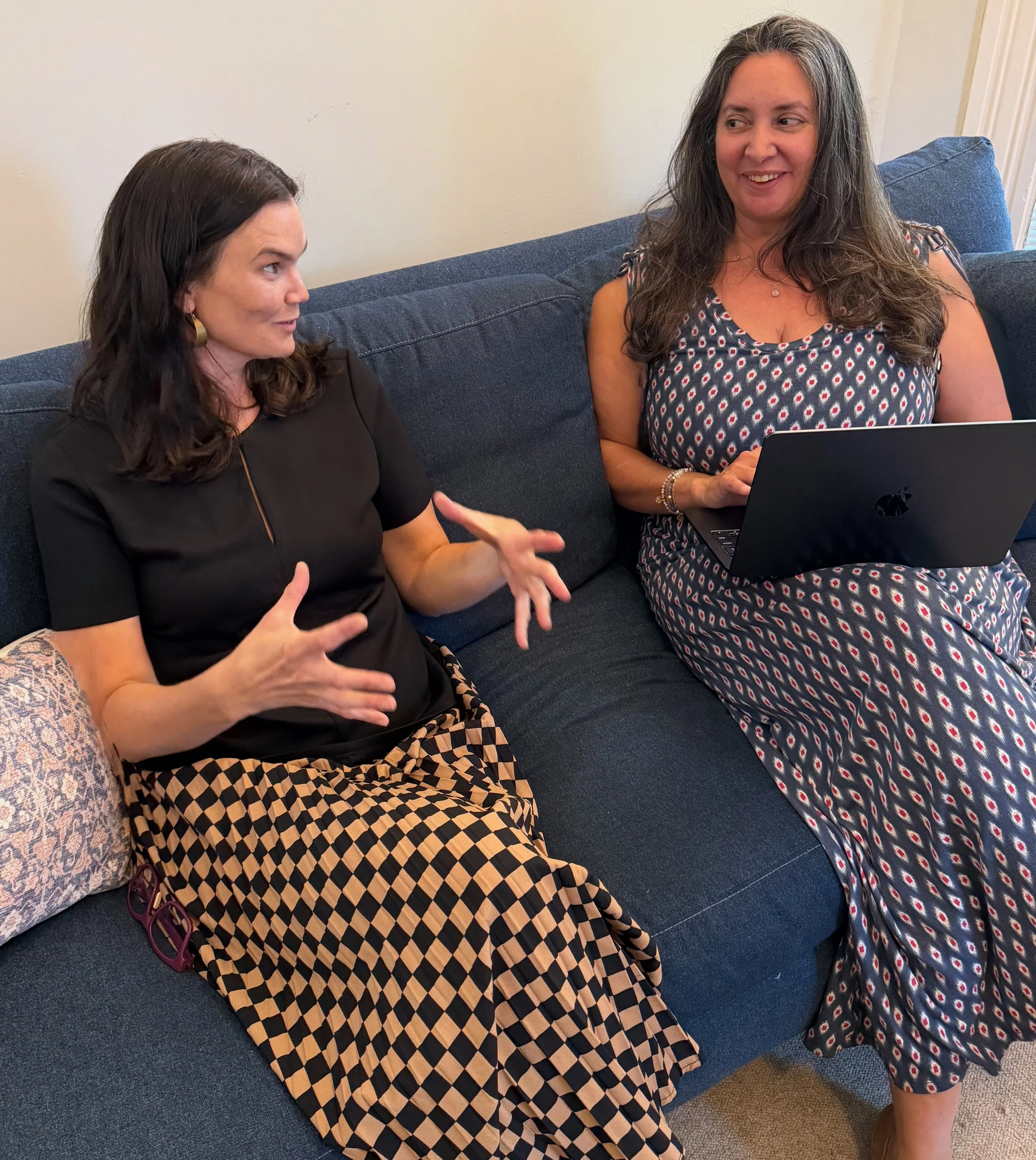 Two women sitting on a blue couch having a conversation. One woman, with dark hair and earrings, is gesturing with her hands. The other woman, with long gray hair, is smiling and holding a laptop.