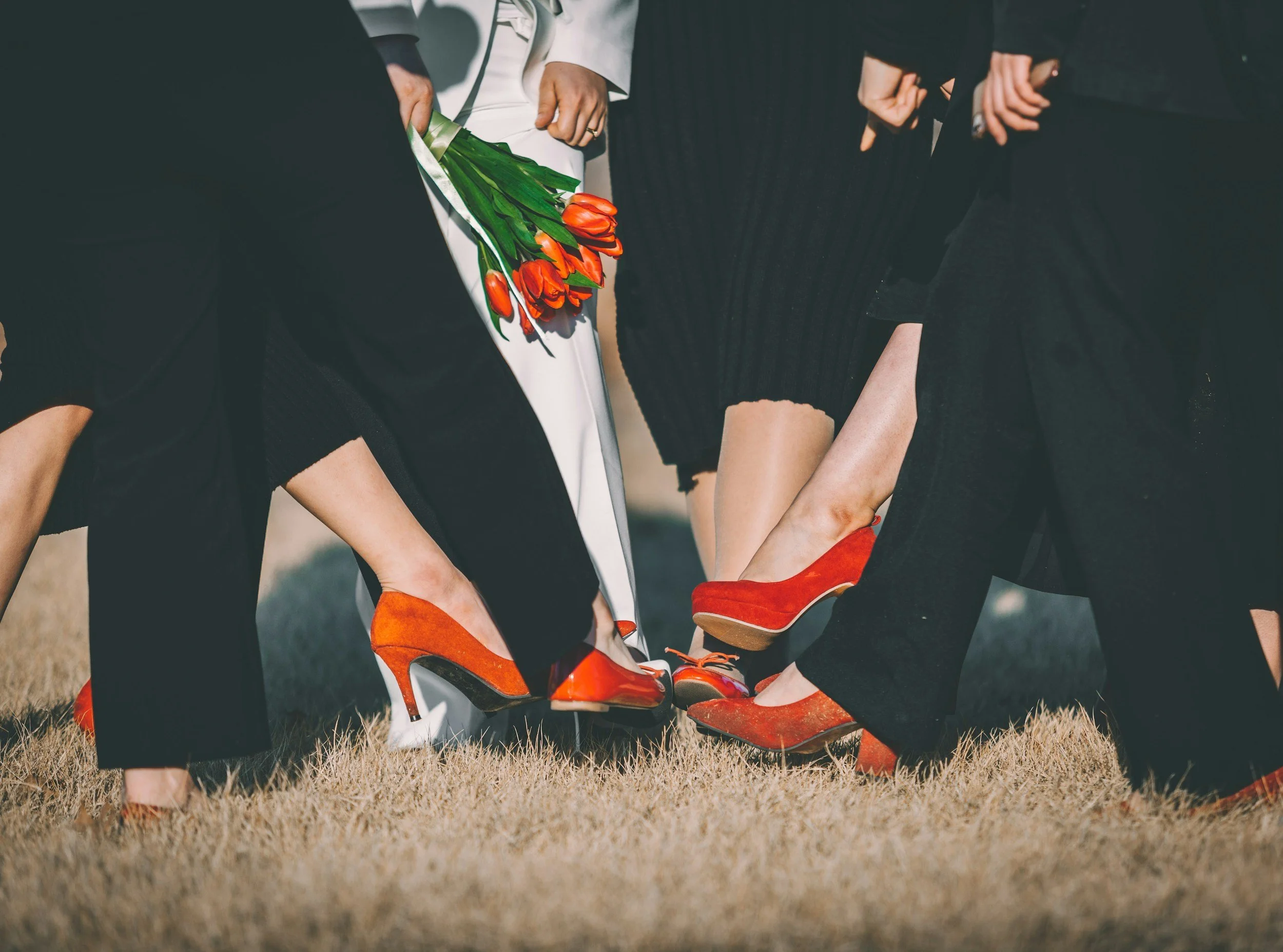 Close-up of people standing in a circle, holding hands, with some wearing red high-heeled shoes and a person holding a bouquet of orange tulips.