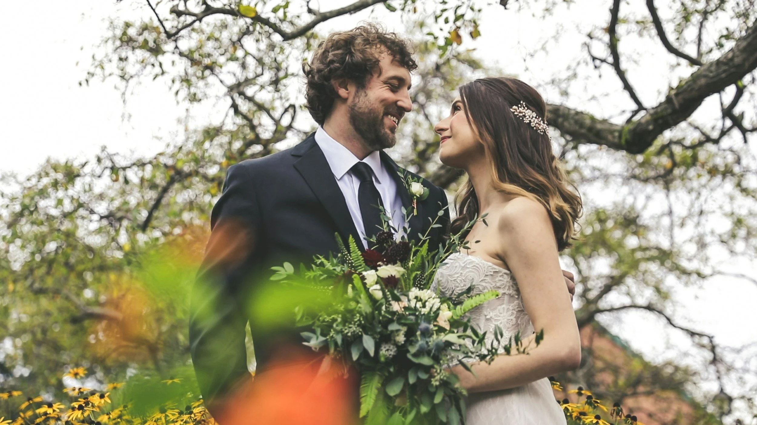 A bride and groom smiling at each other during their wedding outdoors, with trees and cloudy sky in the background.