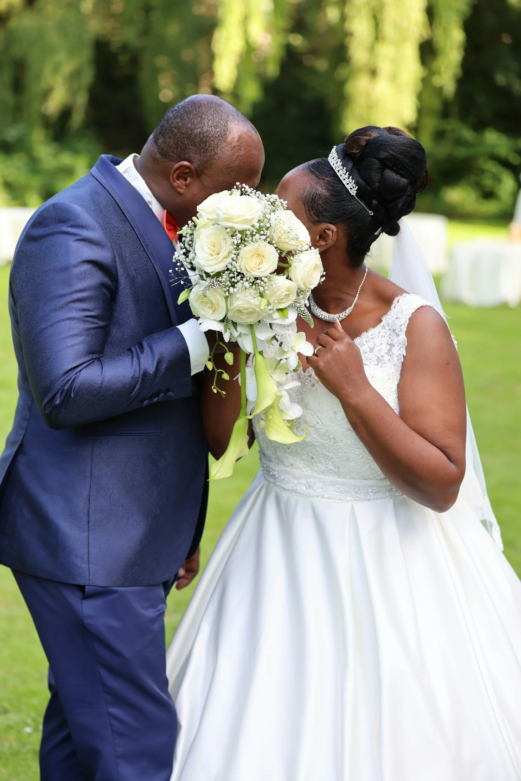 A newlywed couple sharing a kiss outdoors, with the groom in a navy suit and the bride in a white wedding dress holding a bouquet of white roses and greenery.