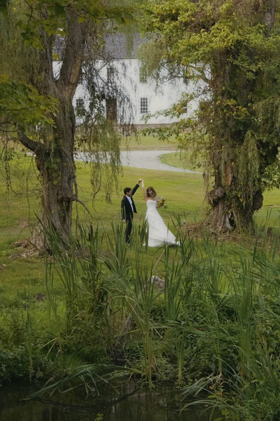 A bride and groom dancing under a large tree in a park, with a pond and a historic white building in the background.
