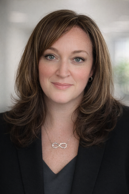 A woman with shoulder-length brown hair, wearing a black blazer and a silver infinity necklace, smiling in an indoor setting with blurred windows in the background.
