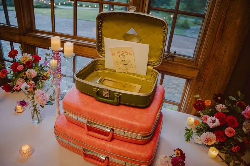 Open vintage suitcase with yellow interior placed on top of two stacked coral-colored suitcases, with a handwritten note and drawing inside the open lid; decorated with flowers and candles on a table near a window.