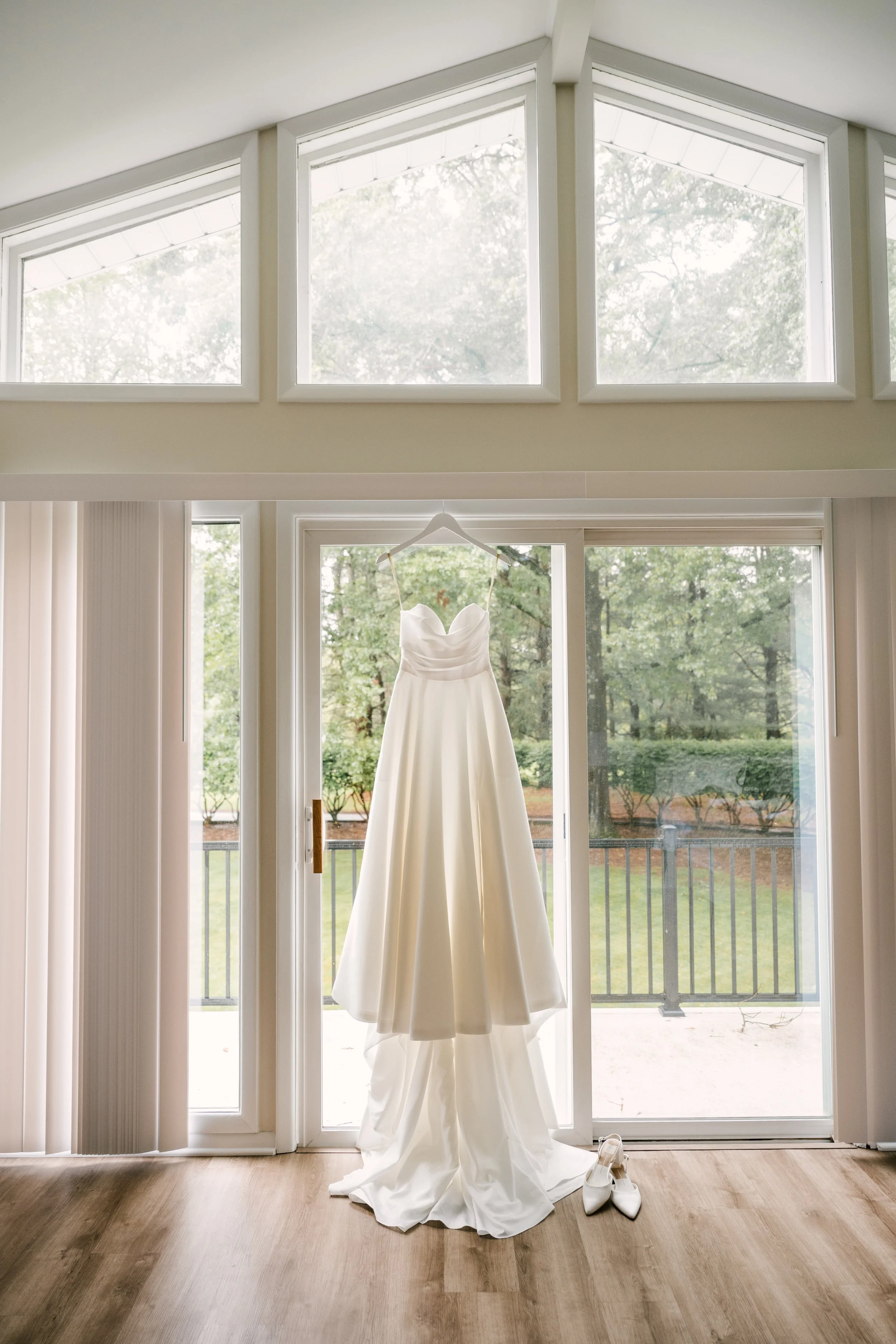 A white wedding dress hanging on a hanger in front of a glass door with a view of trees outside, with a pair of white shoes on the floor.