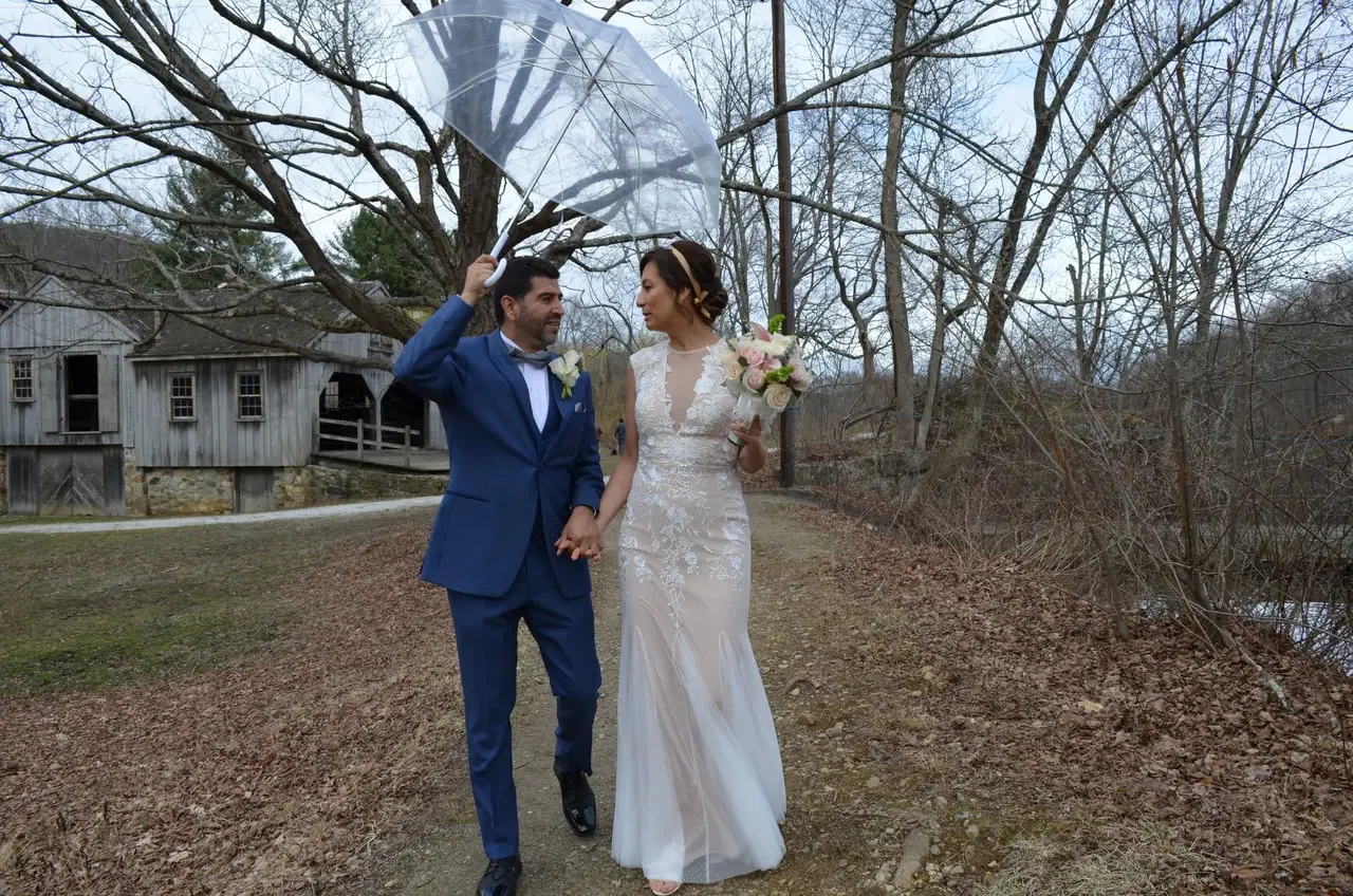 A bride and groom walking outdoors holding hands, with the groom holding a clear umbrella over the bride. The bride is in a white lace wedding dress holding a bouquet of flowers, and the groom is in a blue suit. The background features leafless trees and a rustic wooden building.