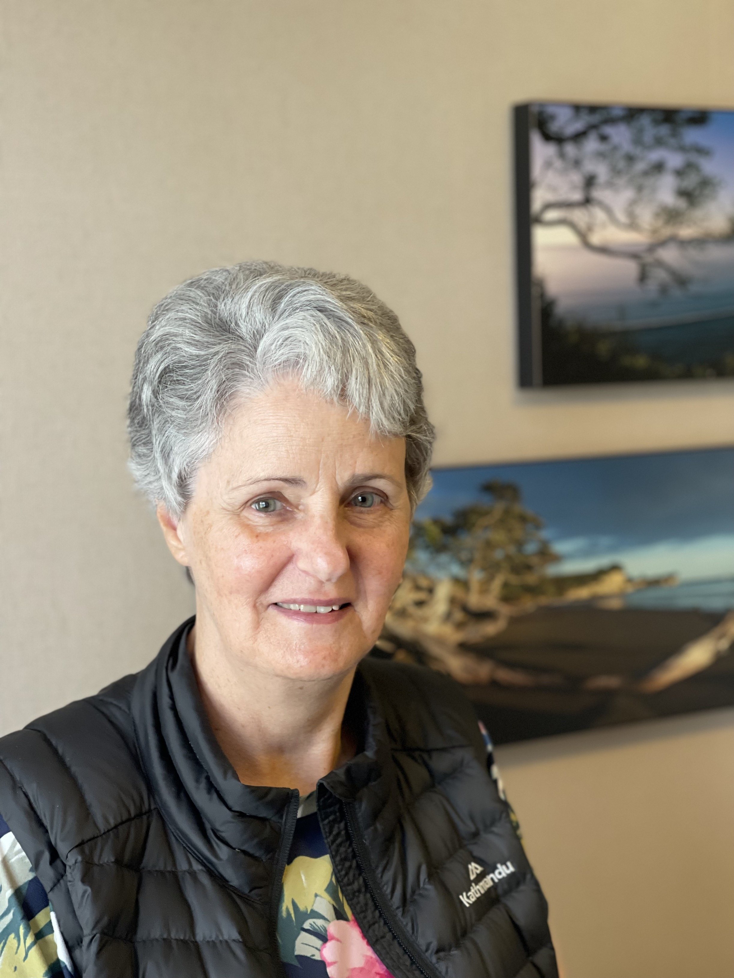 Close-up of a smiling woman with short gray hair, wearing a black puffy jacket with a floral shirt underneath, standing indoors with a beach scene painting in the background.