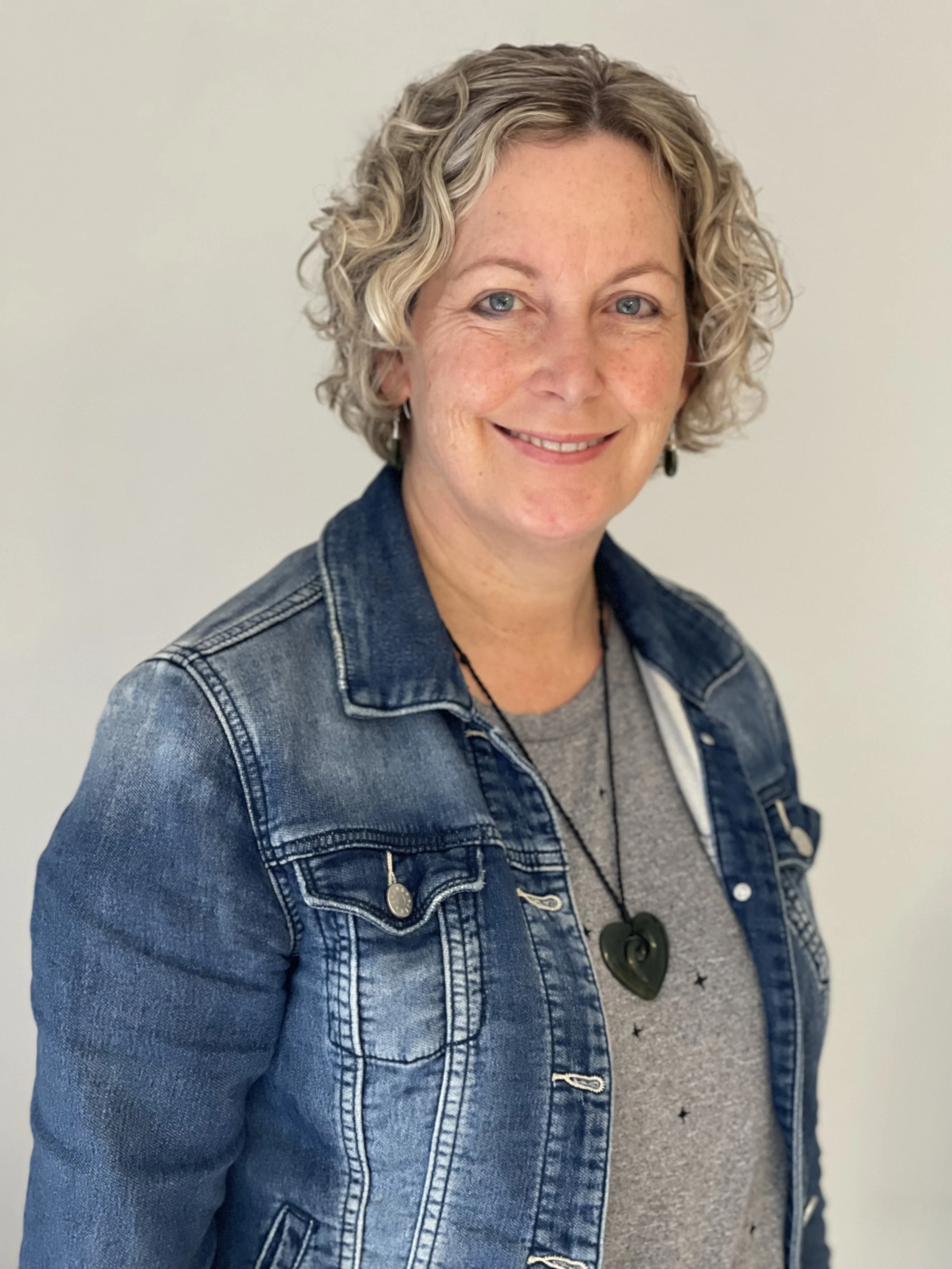 A woman with short curly blonde hair, blue eyes, and light freckles smiling, wearing a denim jacket layered over a gray shirt with star and moon patterns, accessorized with a green swirl-shaped necklace, standing in front of a plain white wall.