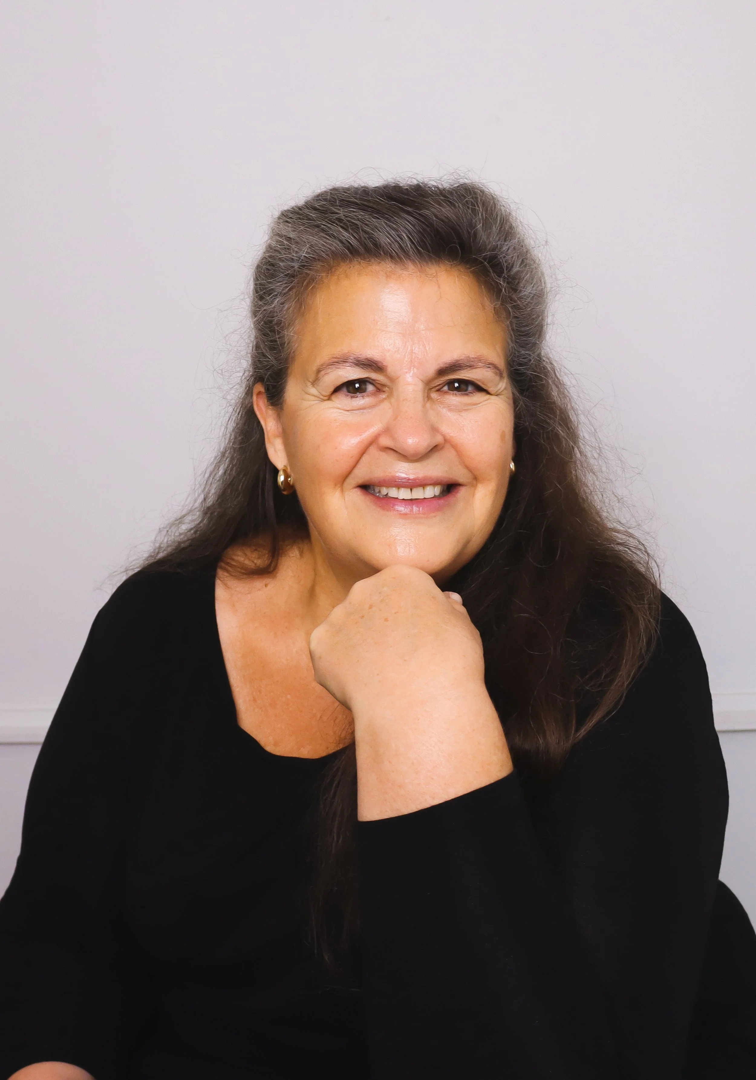 A middle-aged woman with long dark hair, wearing a black top and gold earrings, smiling with her chin resting on her hand, against a plain white background.