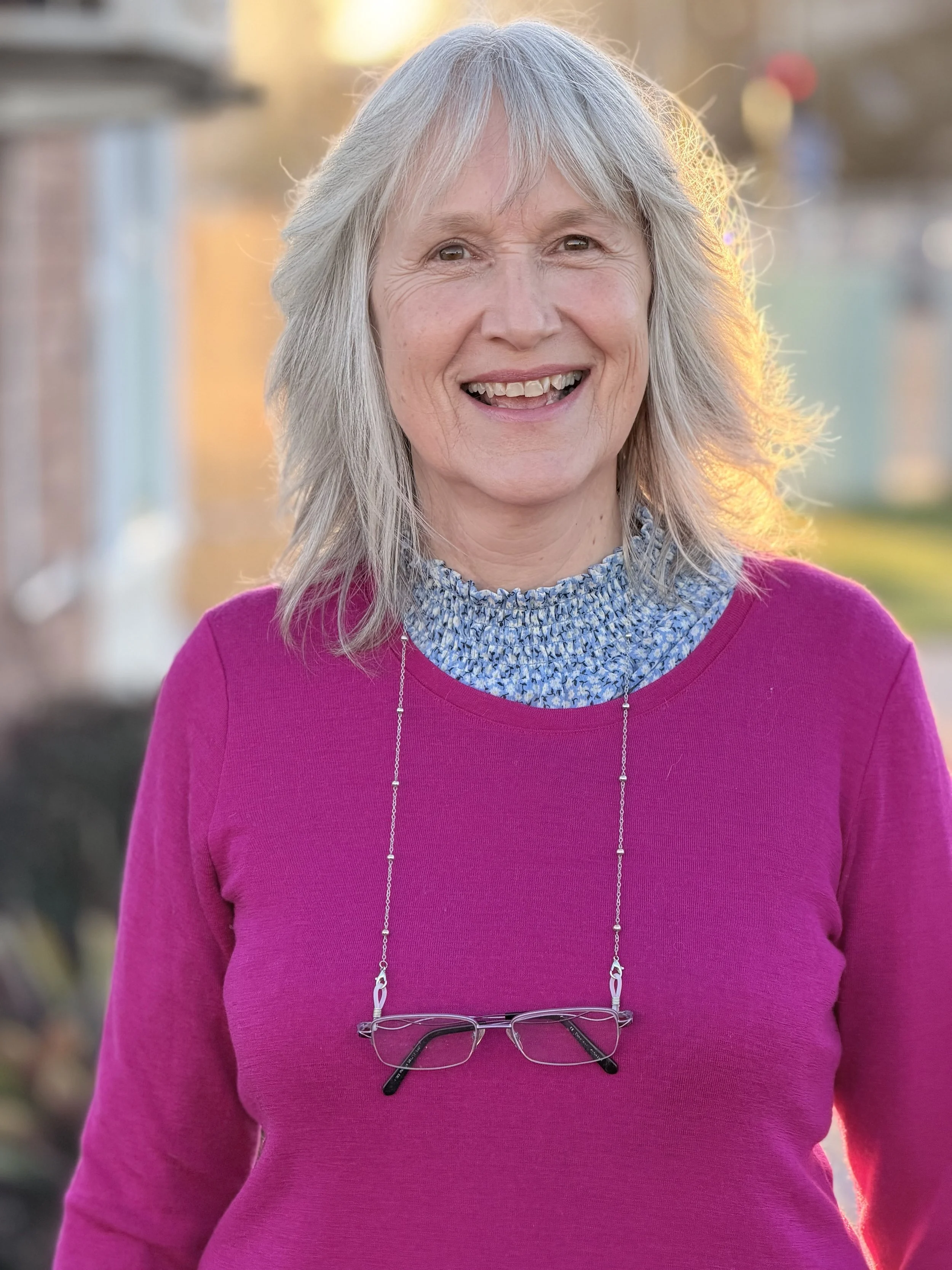 Close-up of a smiling middle-aged woman with gray hair, wearing a pink sweater and a blue patterned shirt, with glasses hanging from her neck, outdoors during sunset.