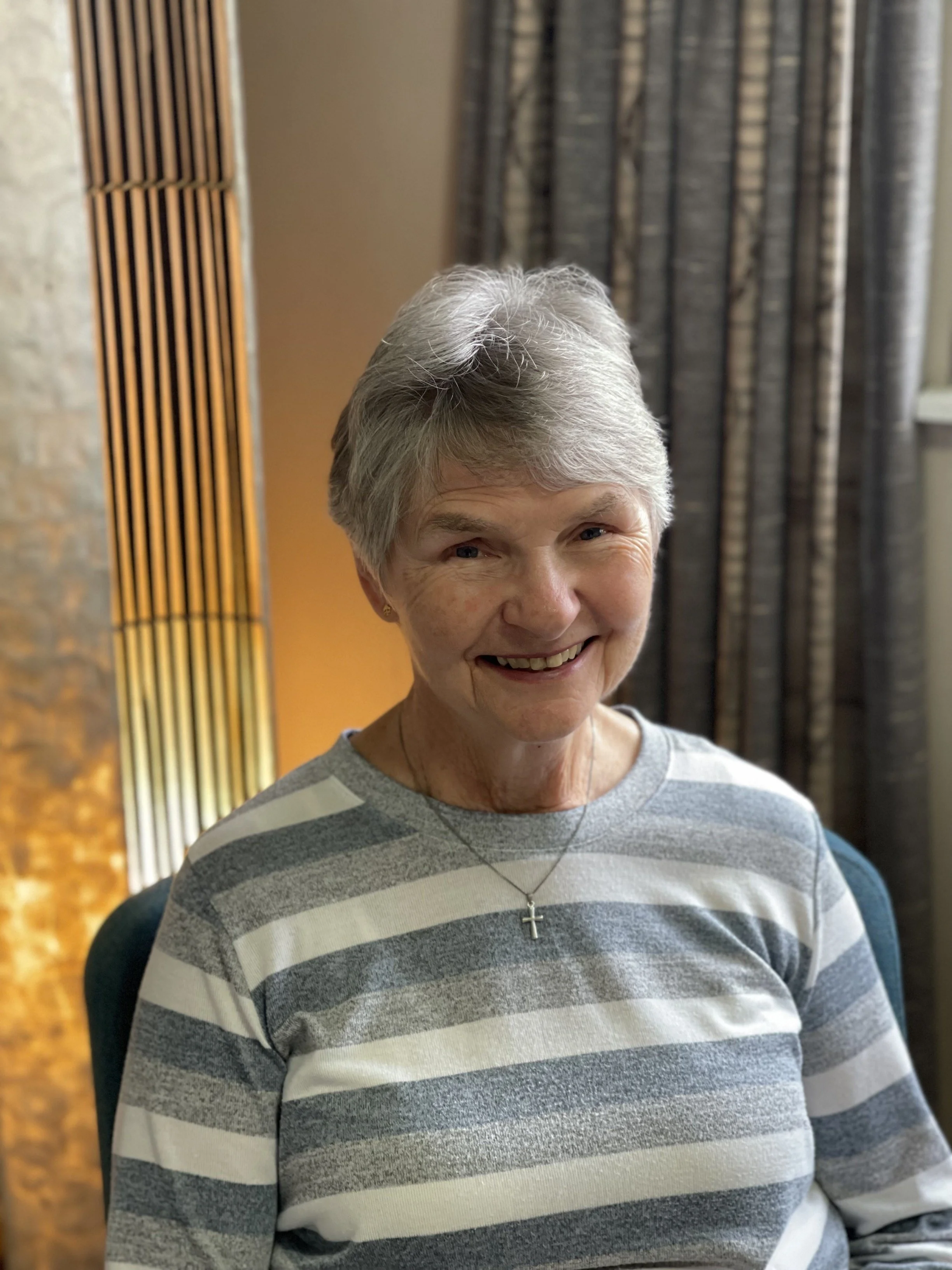 A woman with short gray hair smiling, wearing a gray and white striped shirt and a cross necklace, sitting in front of patterned curtains and a wall with a wooden panel to her left.