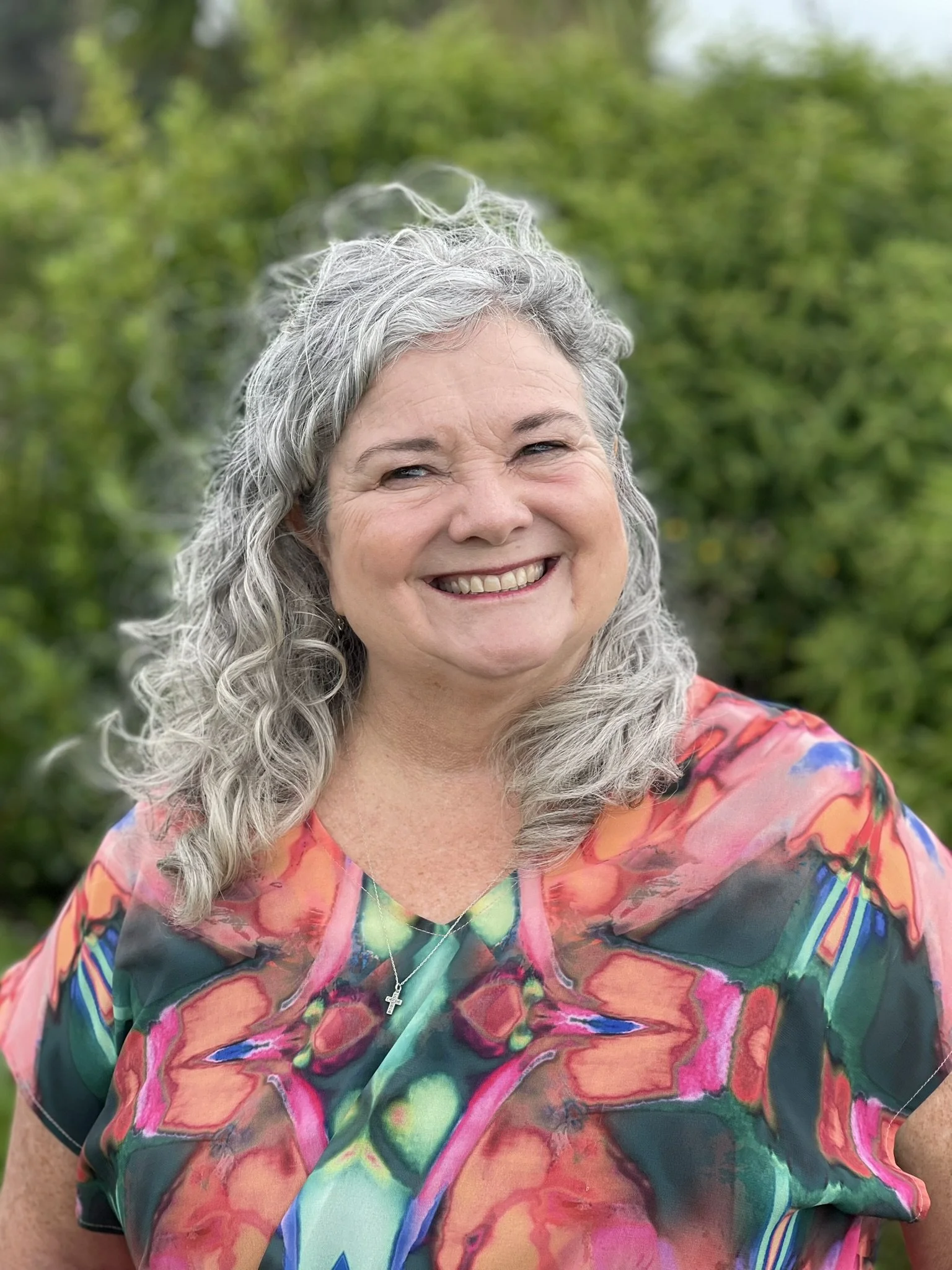 A woman with gray, curly hair smiling outdoors, wearing a colorful, abstract patterned top and a necklace with a cross pendant. Green foliage in the background.