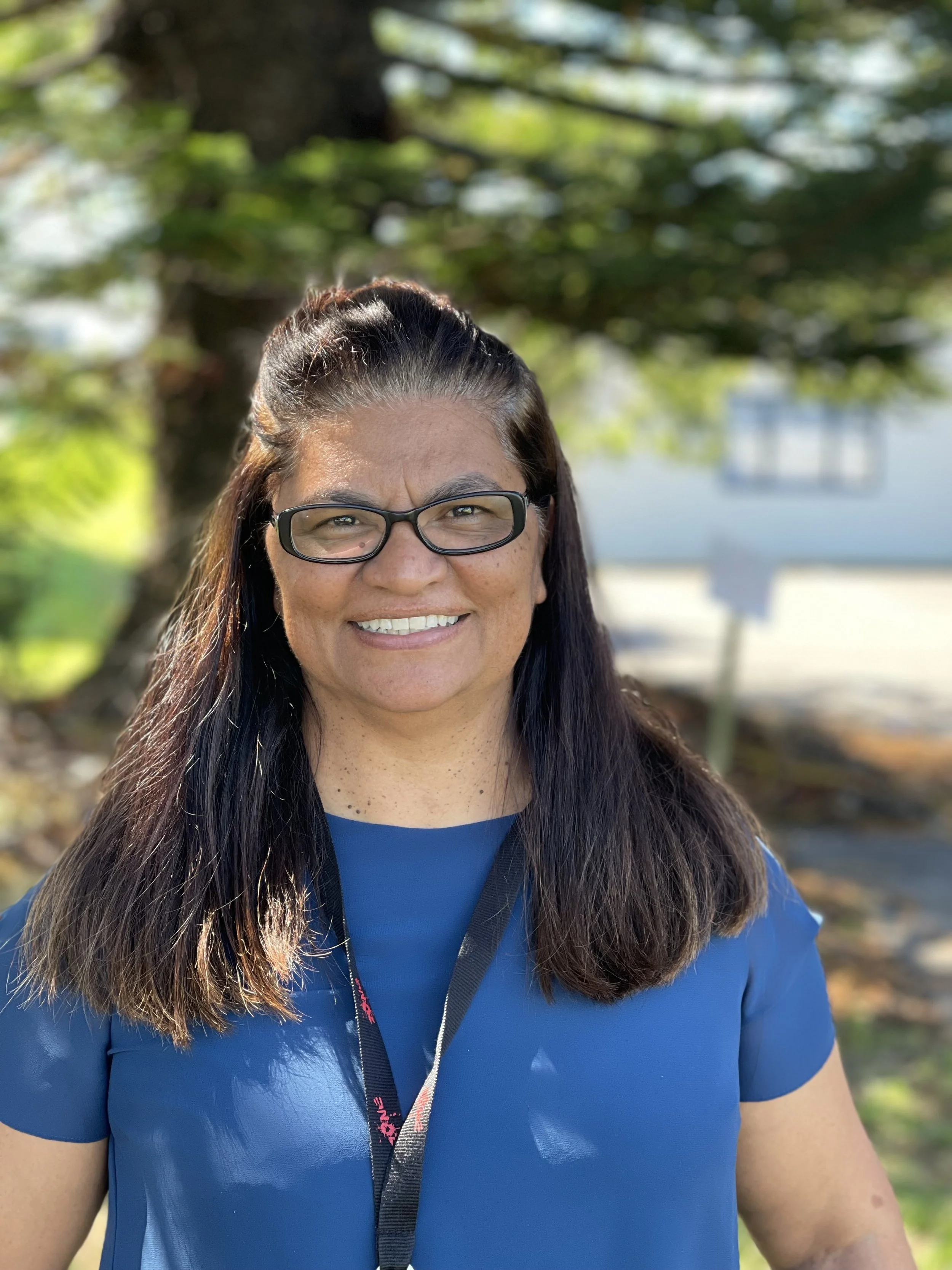 A woman with long dark hair, glasses, and a big smile, wearing a blue athletic shirt, outdoors with green trees and blurred background.