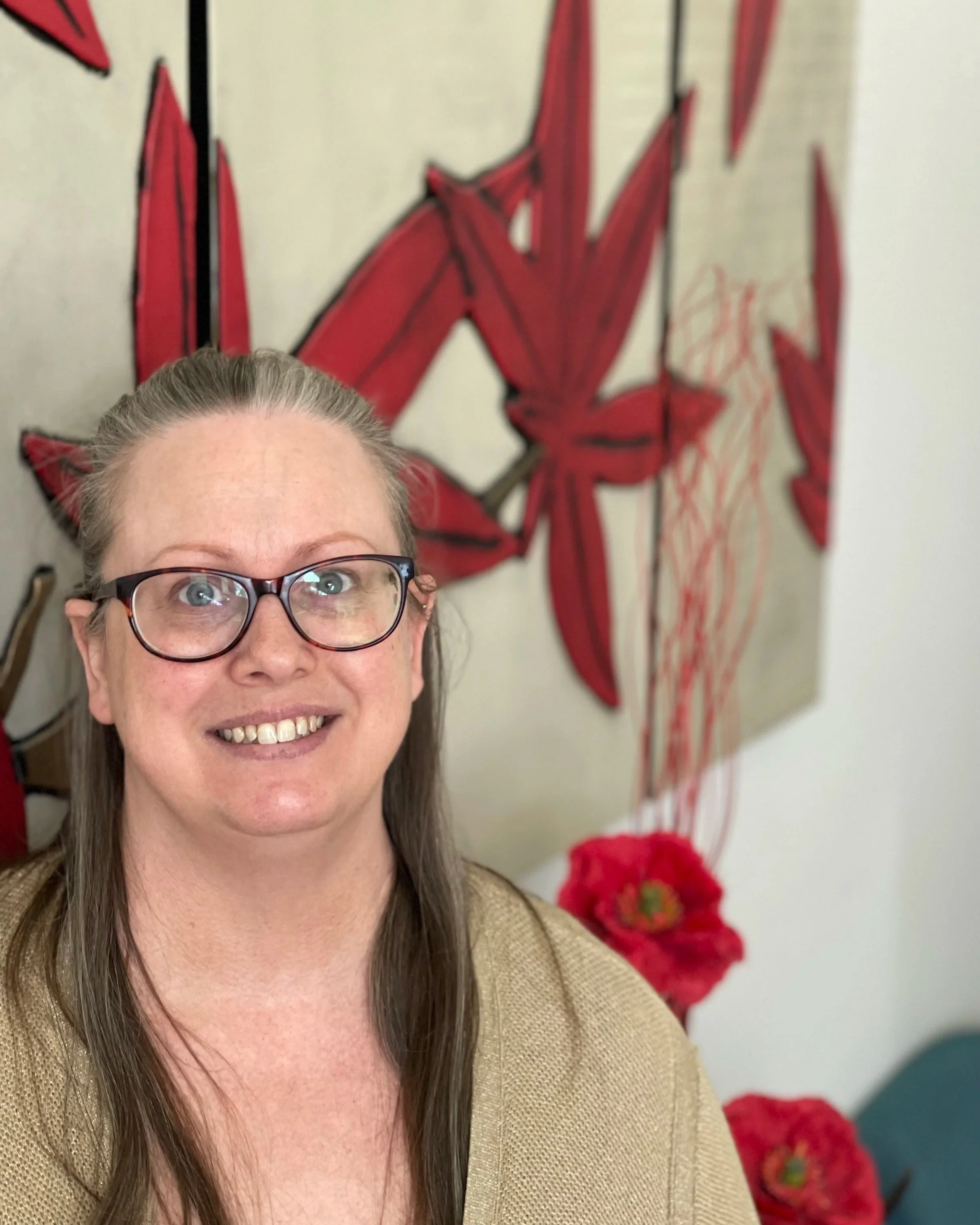 A woman with glasses smiling, with red flowers and a red and white abstract wall art in the background.