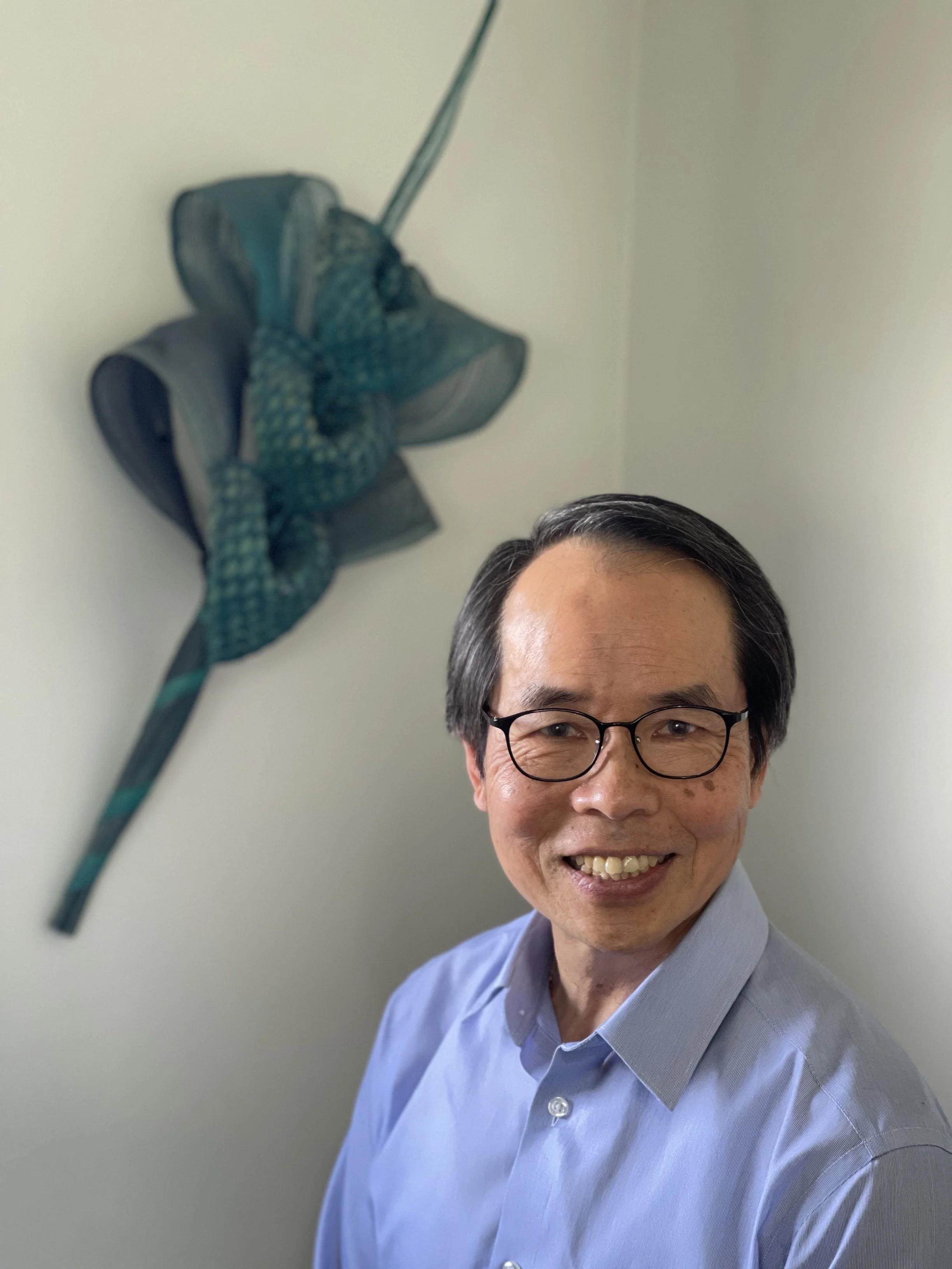 A man with glasses smiling, wearing a blue dress shirt, standing in front of a plain light-colored wall, with a slightly blurred dishtowel or cloth hanging on the wall behind him.