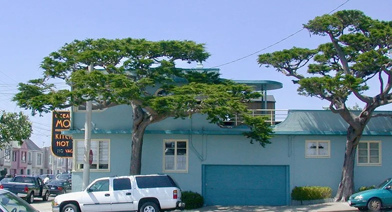 A light blue building with tall, flat-topped trees in front, parked cars, and a sign that reads 'CEAN' and 'MO' visible on the left.
