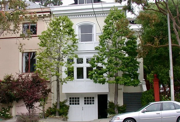 White multi-story house with large windows, surrounded by green trees and a parked silver car in front.