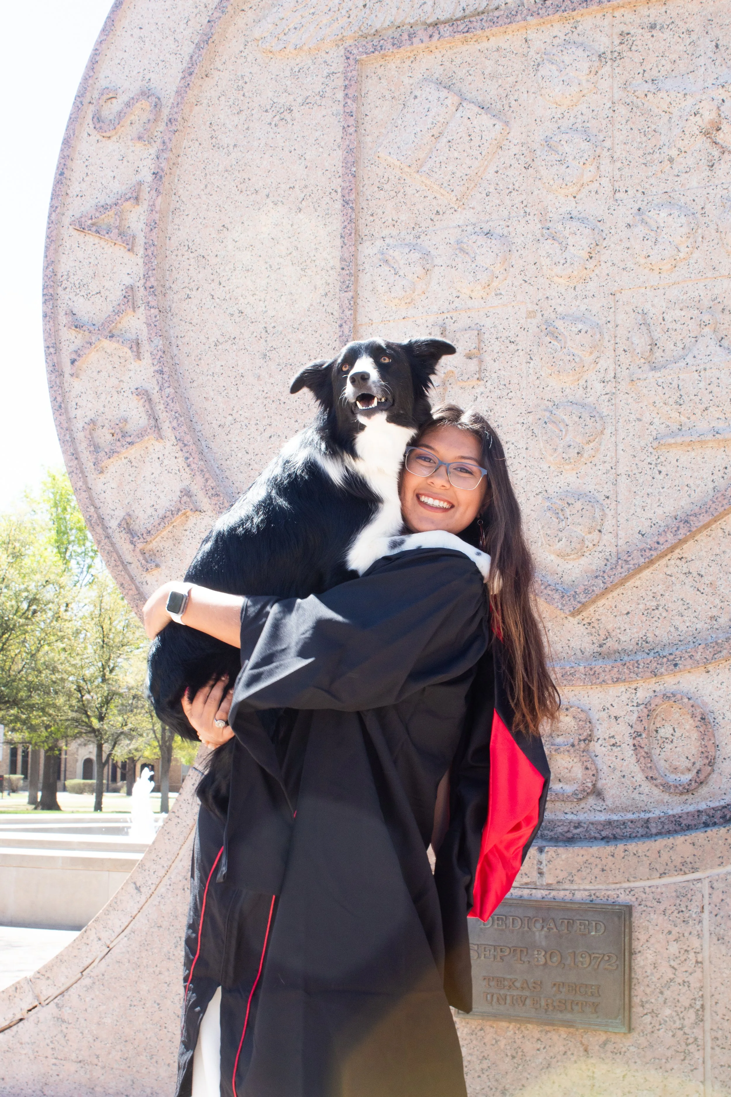 A young woman with long brown hair and glasses holds a black and white dog in front of a large stone monument with engraved text. The woman is smiling, wearing a black jacket with red accents, and the dog has an alert expression. The background features trees and a clear sky.