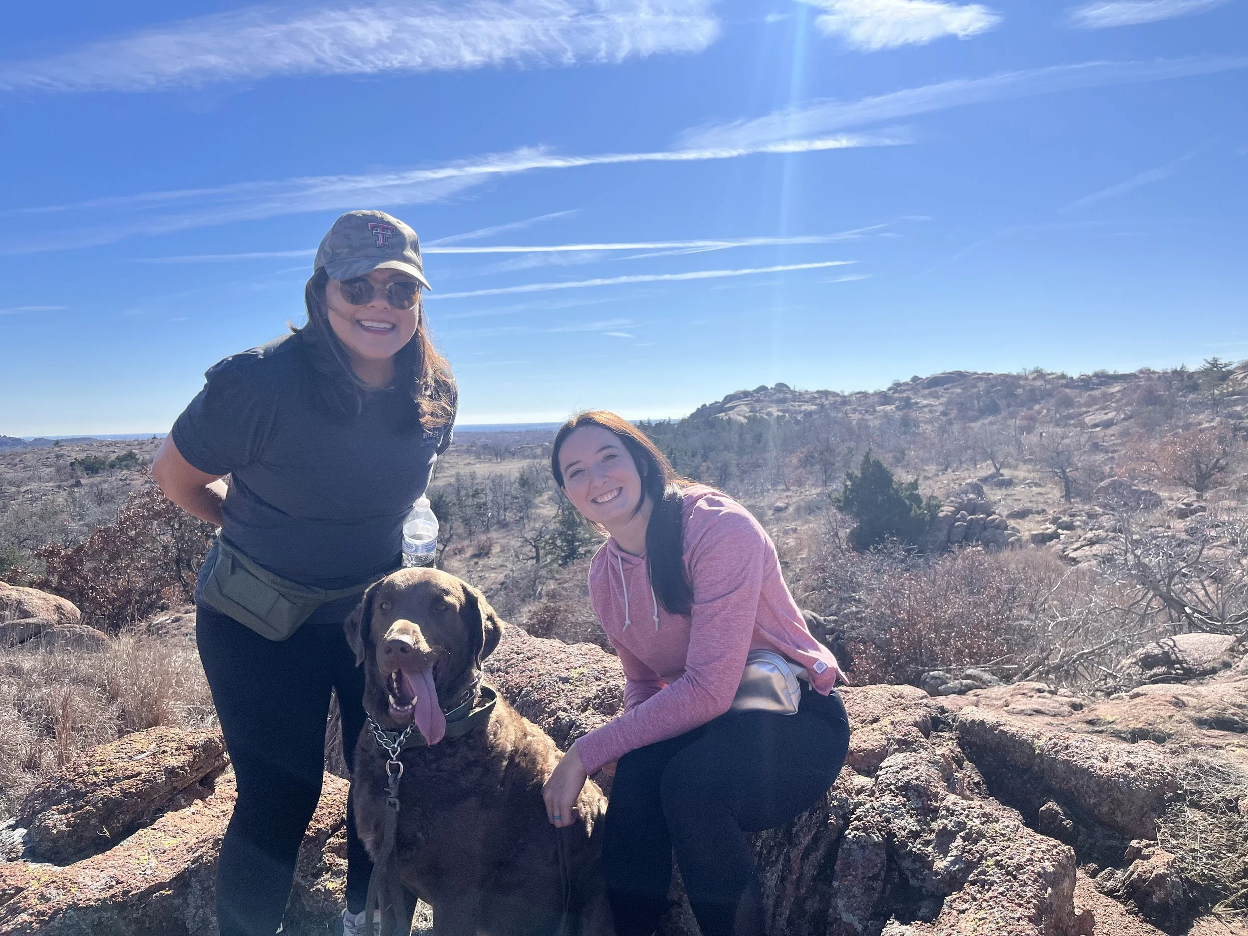 Two women and a large dog hiking in a rocky, dry landscape with sparse vegetation, under a bright blue sky with wispy clouds.