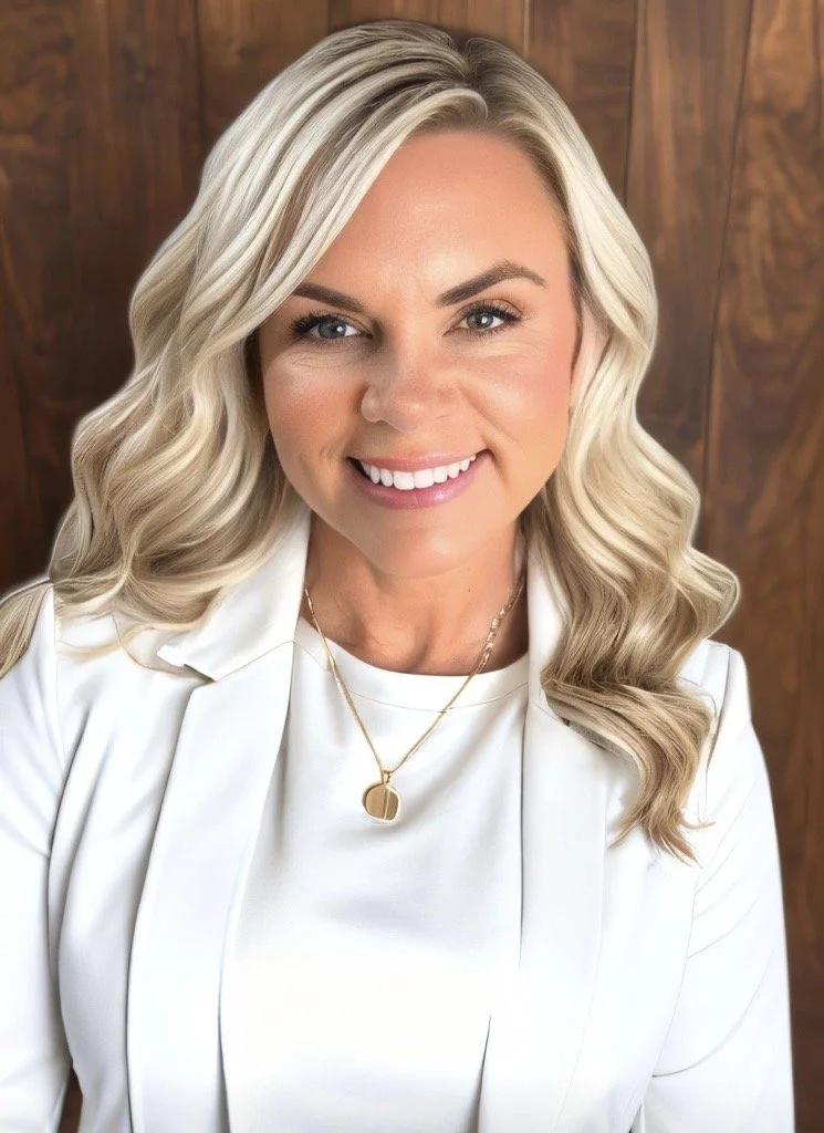 A woman with blonde, wavy hair smiling at the camera, wearing a white blazer and gold jewelry, in front of a wooden background.