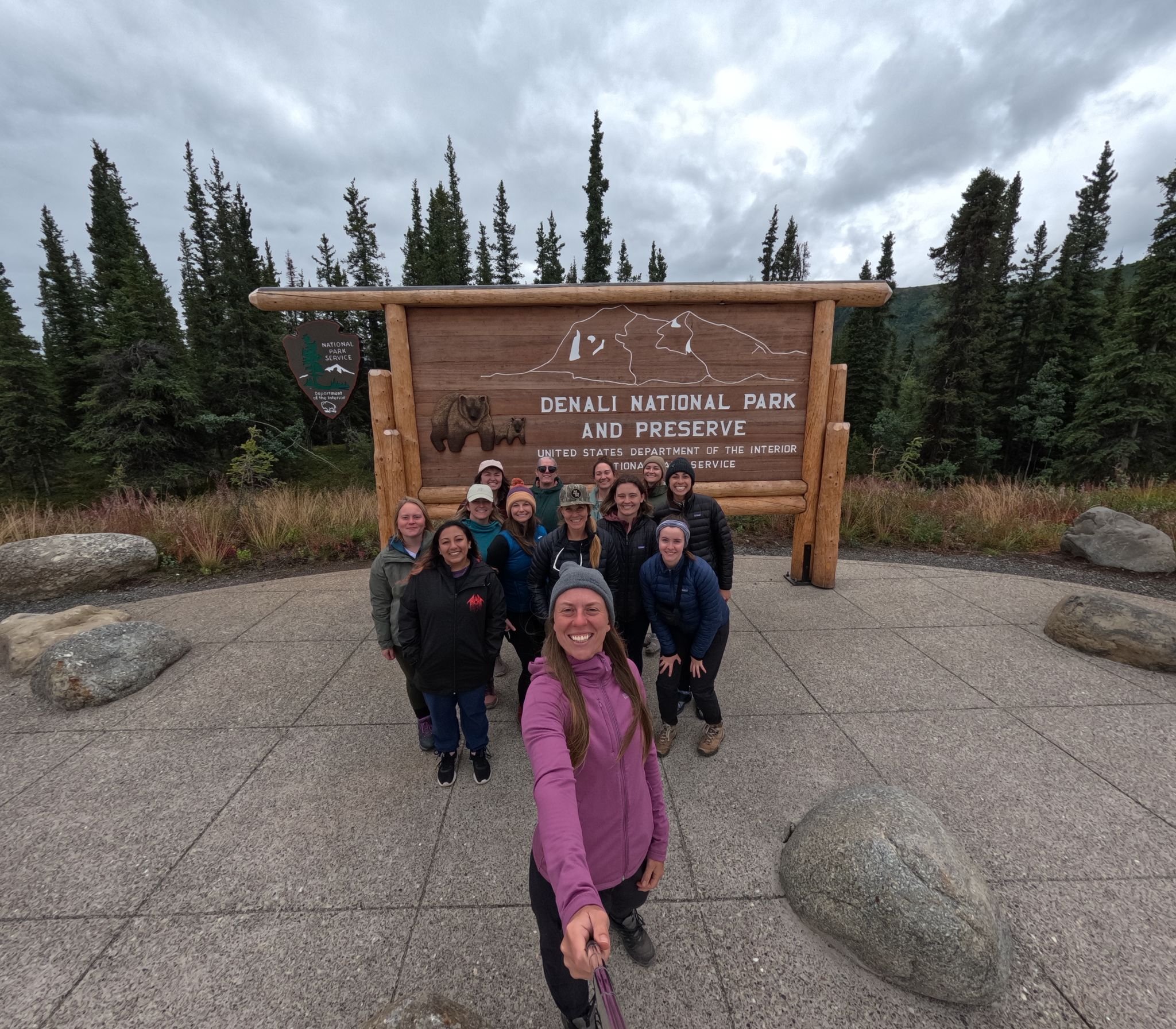Group of smiling people taking a selfie in front of a Denali National Park sign with trees and cloudy sky in the background.