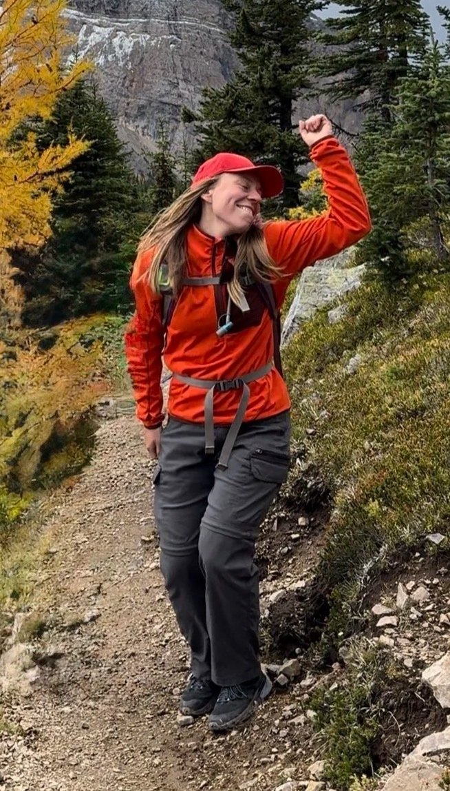 A woman hiking on a trail in a forest with fall foliage, wearing a red jacket, red cap, and carrying a backpack, with a big smile and raising her right arm in victory.