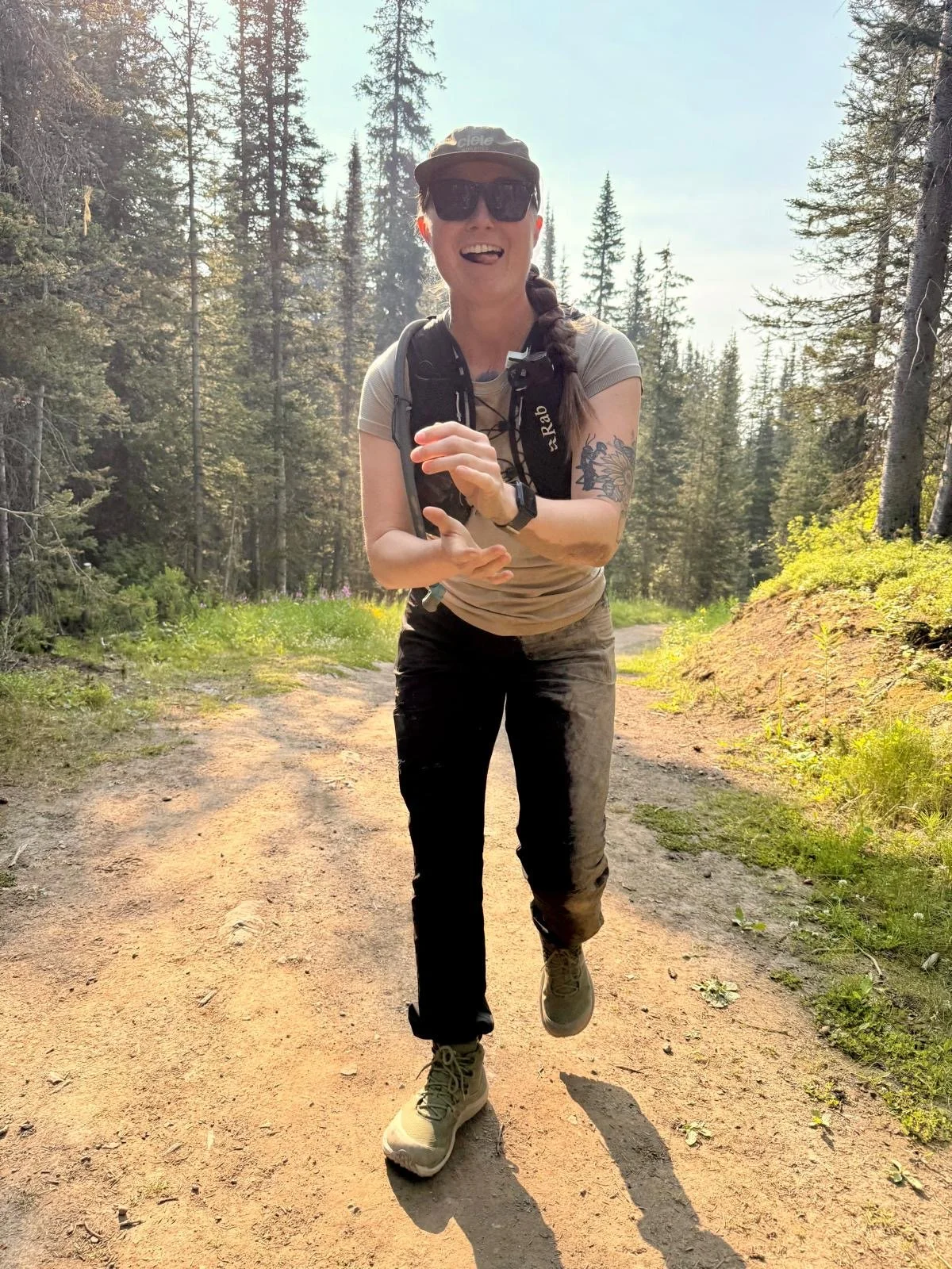 A woman with a braid, wearing sunglasses, a cap, a light-colored t-shirt, and black pants, is jogging on a dirt trail in a forest. She is smiling and appears happy, with tall trees and green foliage in the background.