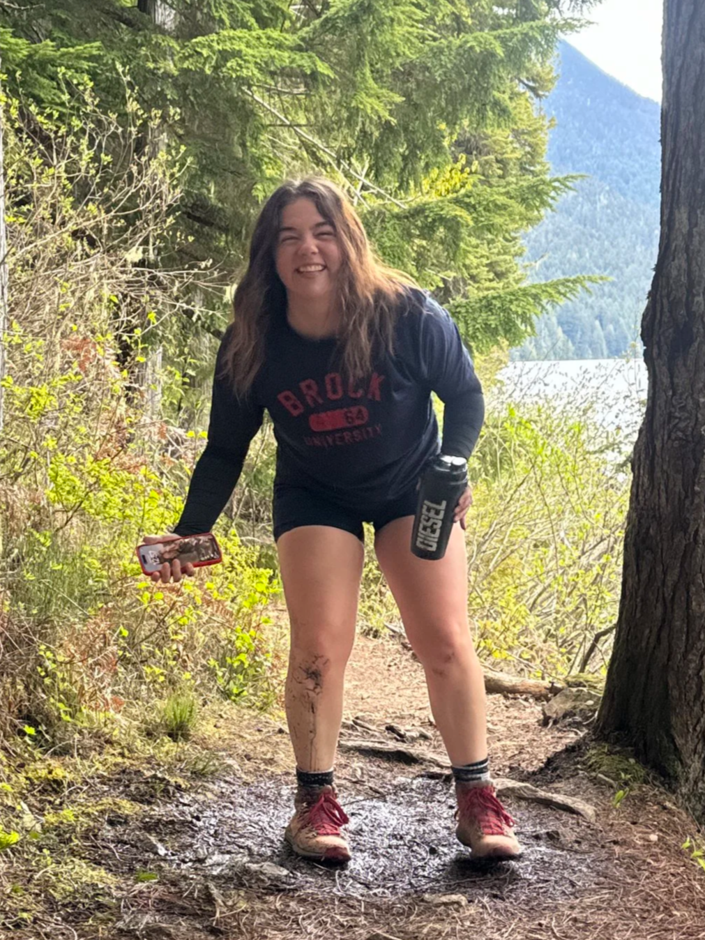A woman with long brown hair smiling, wearing a Brock University t-shirt, shorts, and muddy trail running shoes, standing on a dirt trail in a forested area near a lake with mountains in the background.