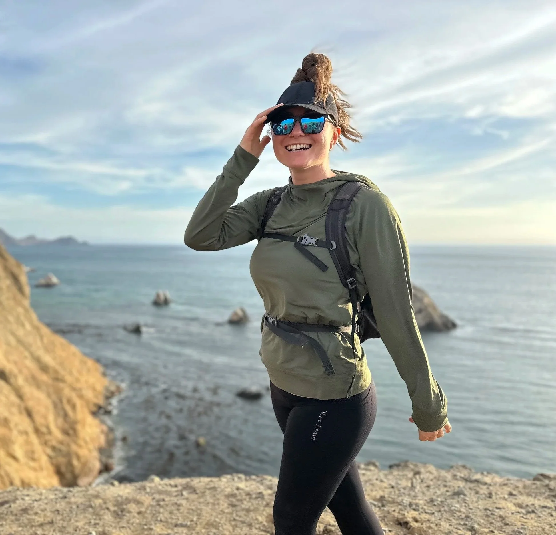 A woman dressed in athletic gear standing on a rocky beach near the ocean, smiling, wearing sunglasses, a black cap, and a backpack, with a cloudy sky background.