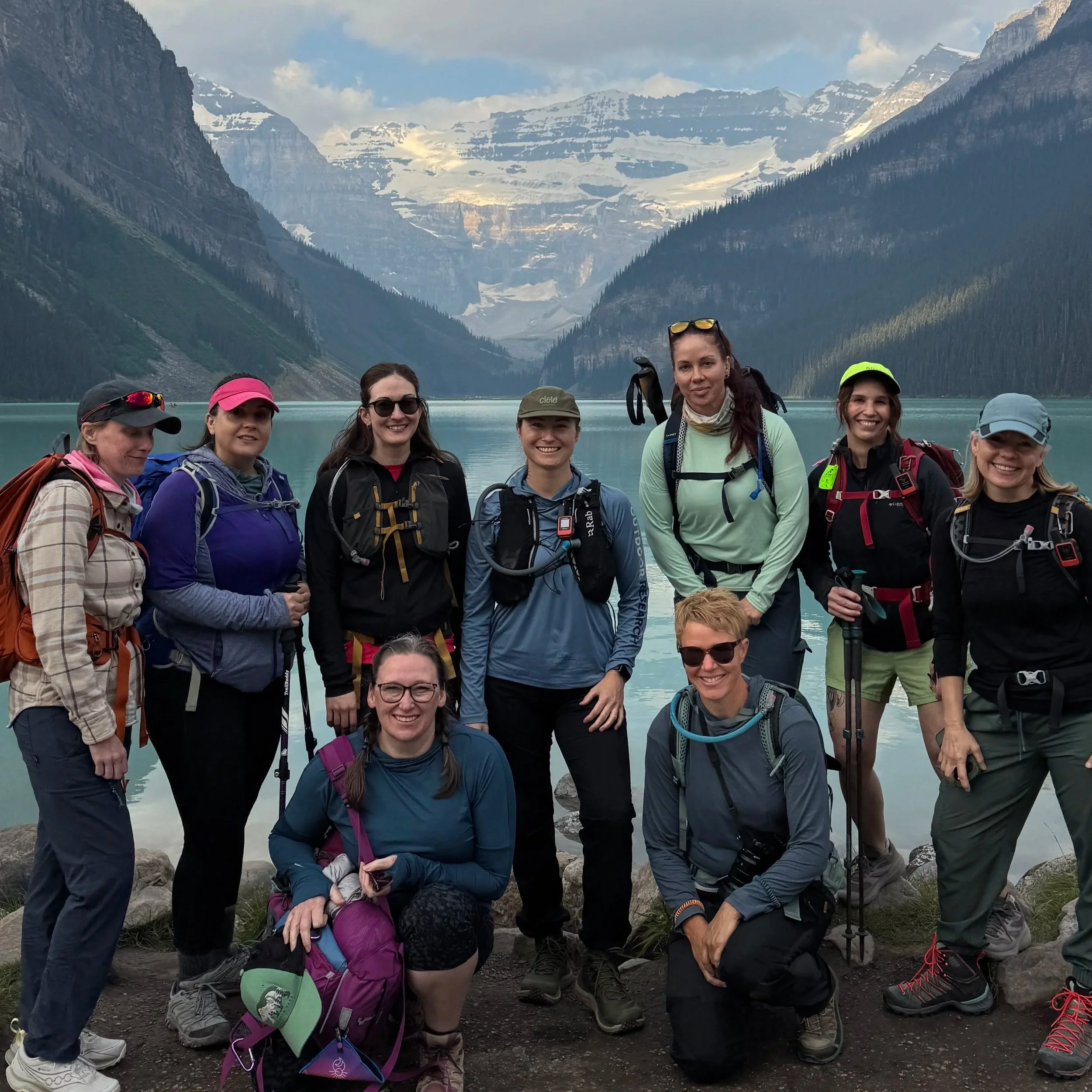 A group of hikers standing together on a rocky shoreline with a mountain lake and snow-capped mountains in the background, under cloudy skies.