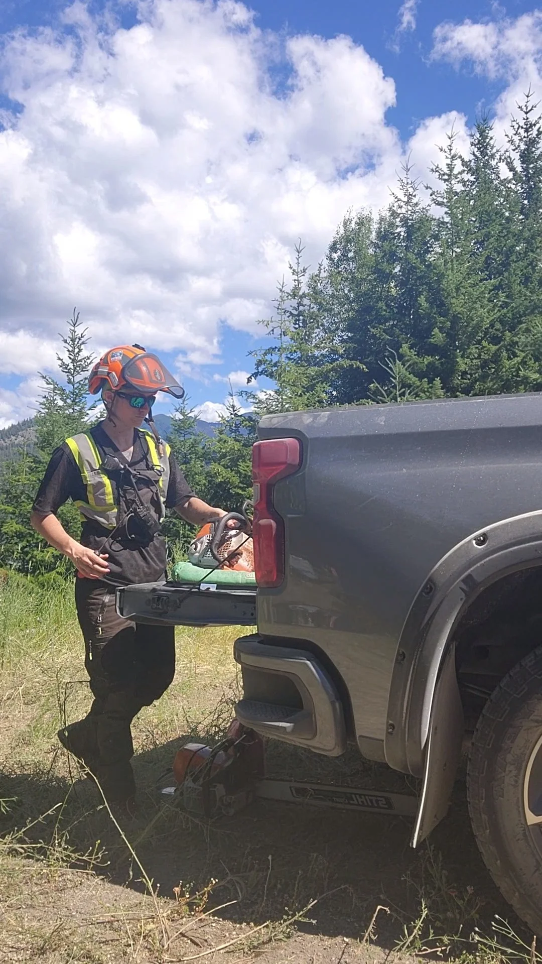 Person in safety gear standing next to a pickup truck with the tailgate down, outdoors with trees and a partly cloudy sky in the background.