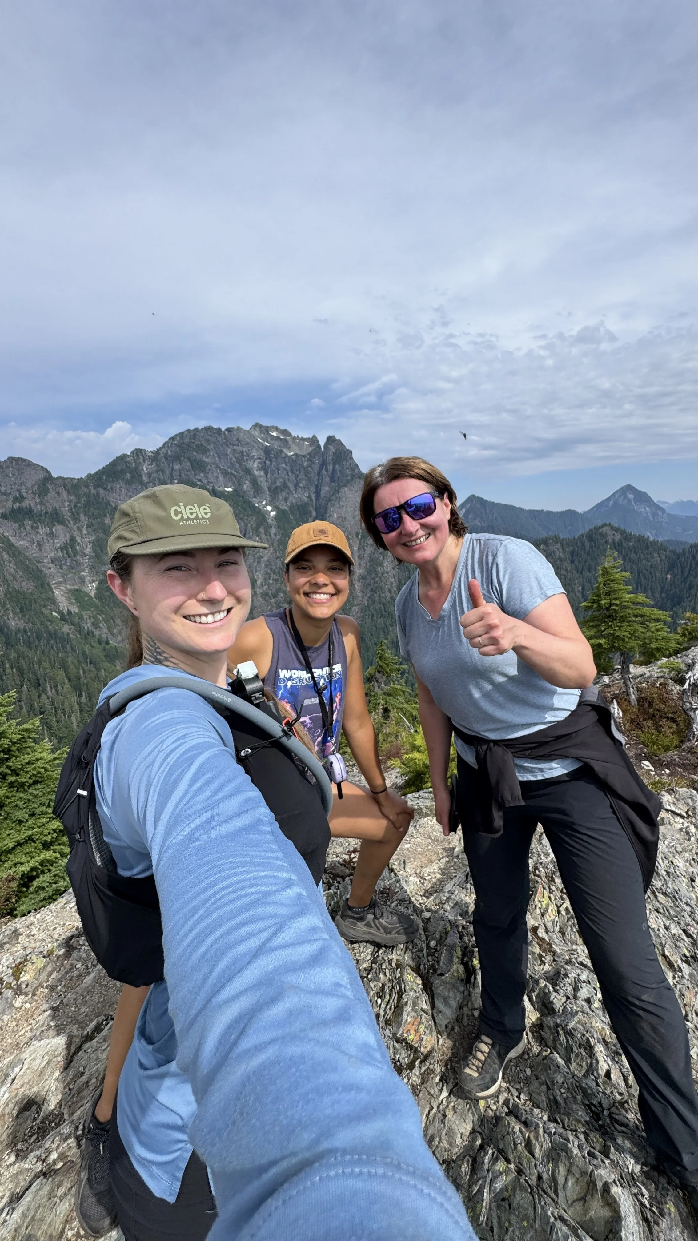 Three women smiling and posing for a selfie on a mountain trail with rocky terrain, dense green forest, and mountain peaks in the background, under a partly cloudy sky.
