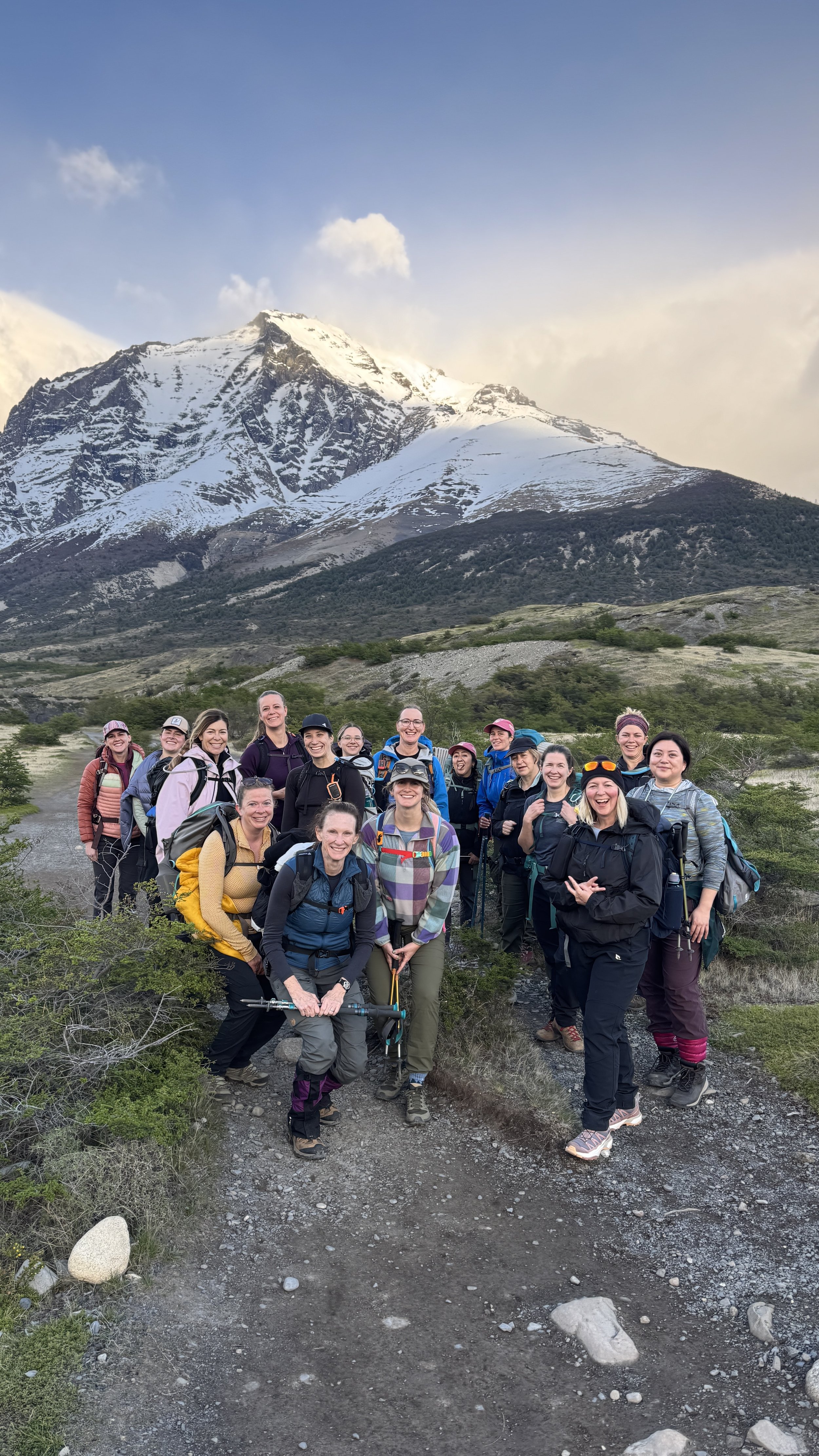 A group of hikers with backpacks and outdoor gear standing on a trail with snow-capped mountain in the background.