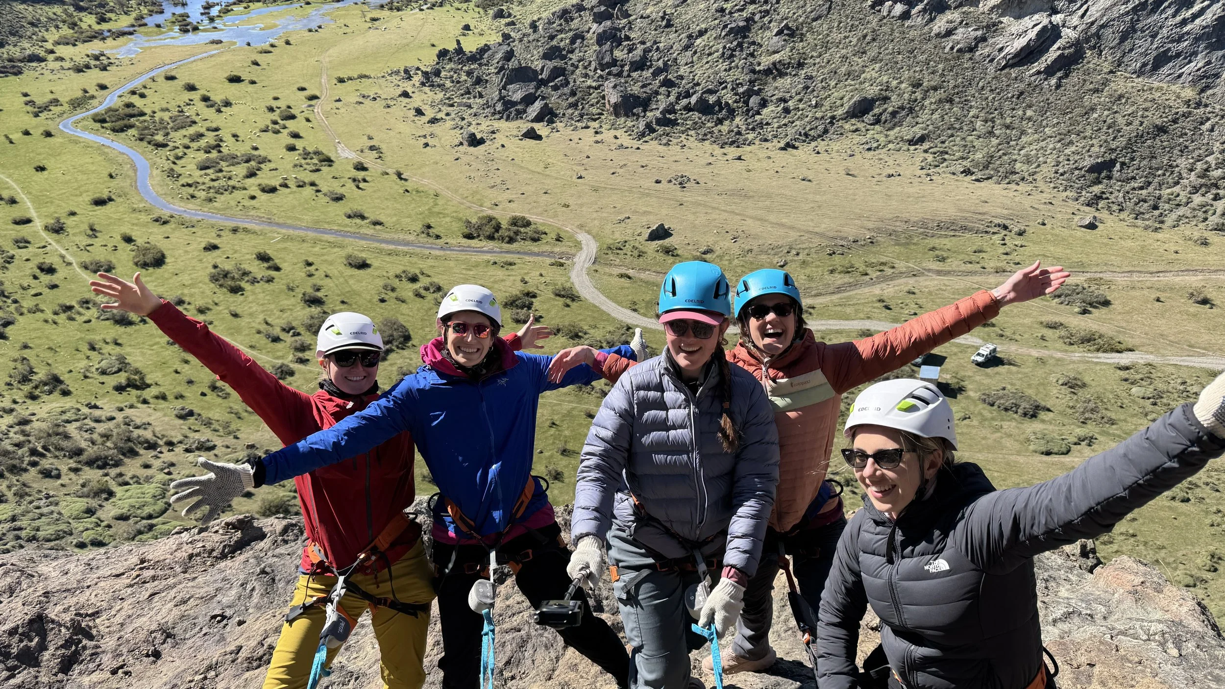 Group of five women in outdoor climbing gear smiling and posing with arms outstretched on a mountain ridge with a valley, river, and rocky hillside in the background.