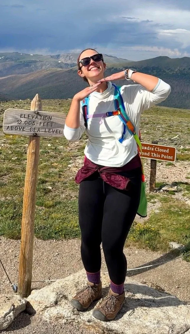 Woman smiling and posing with a playful gesture at a mountain summit, wearing hiking gear, sunglasses, and a backpack, with a sign indicating an elevation of 12,006 feet above sea level in a rugged mountainous landscape.