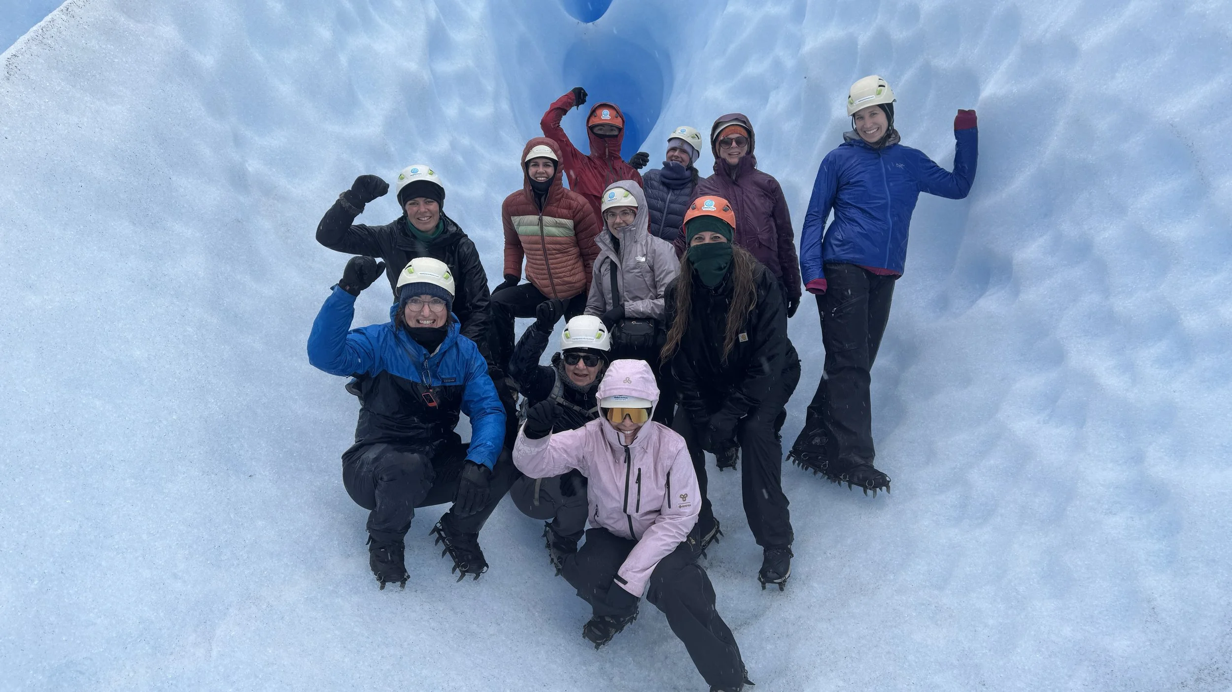 Group of nine people in winter gear and helmets posing inside a glacier ice tunnel, smiling and making fist gestures.