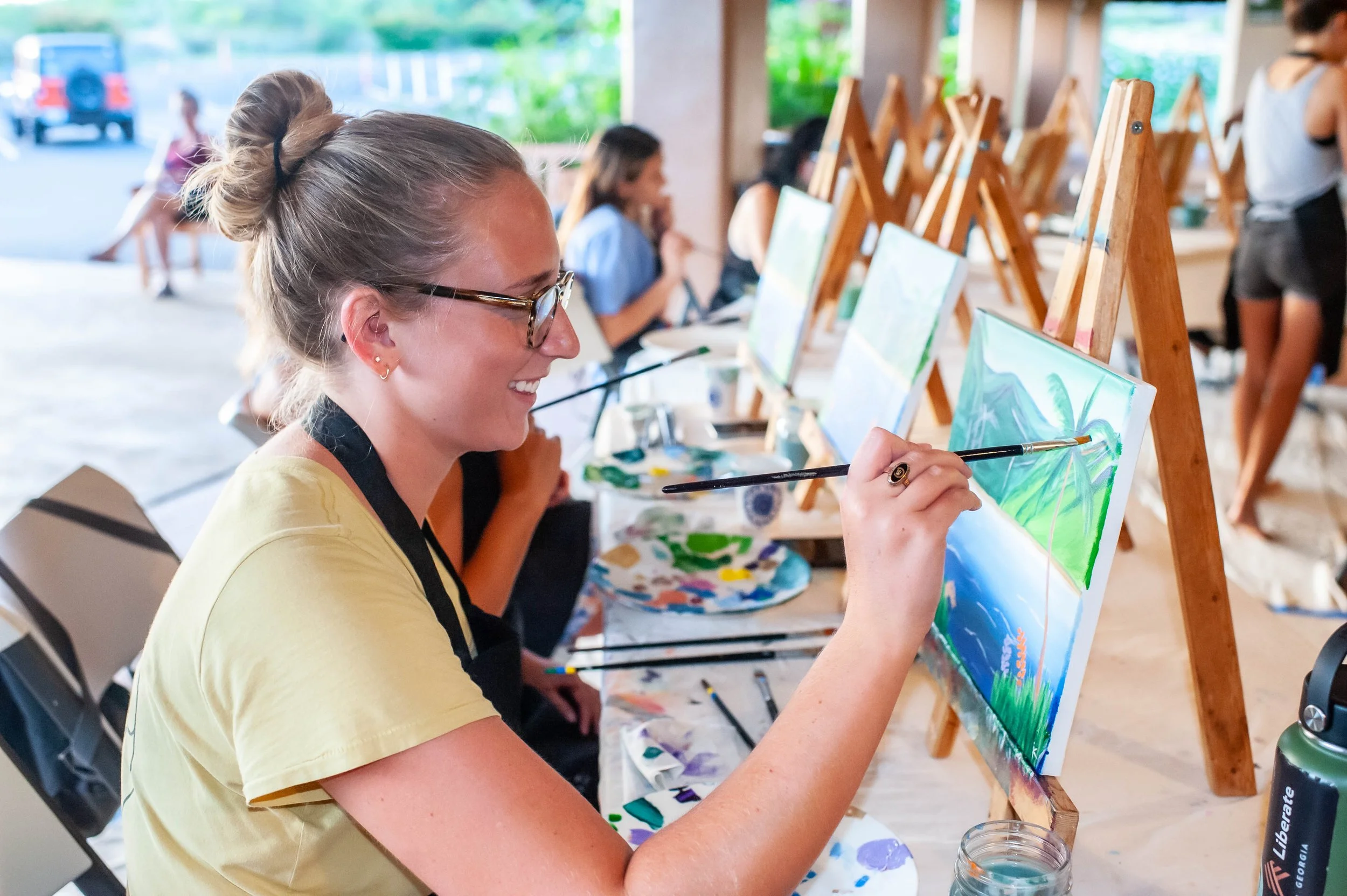 A woman painting a landscape on a canvas at an art class, seated at a table with other students also painting, in a well-lit room with large windowsPainting class on kauai instructed