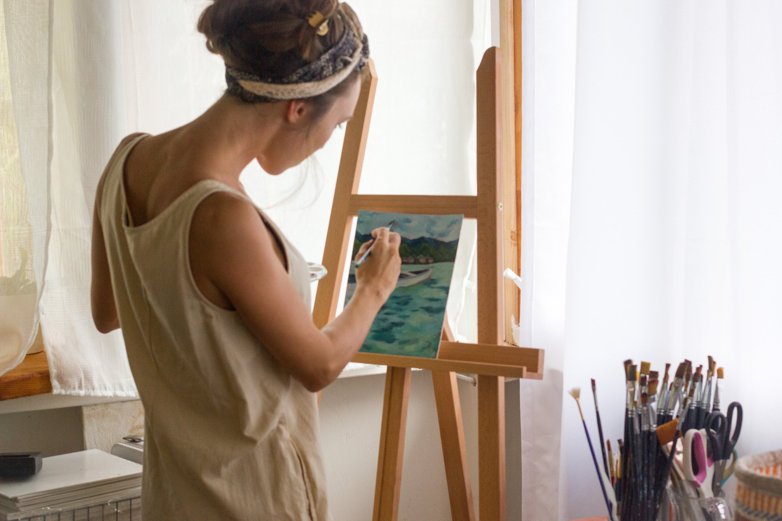 A woman painting a landscape on a canvas placed on an easel. She is standing indoors near a window with sheer white curtains, wearing a sleeveless top and a headwrap. Art supplies are visible nearby.