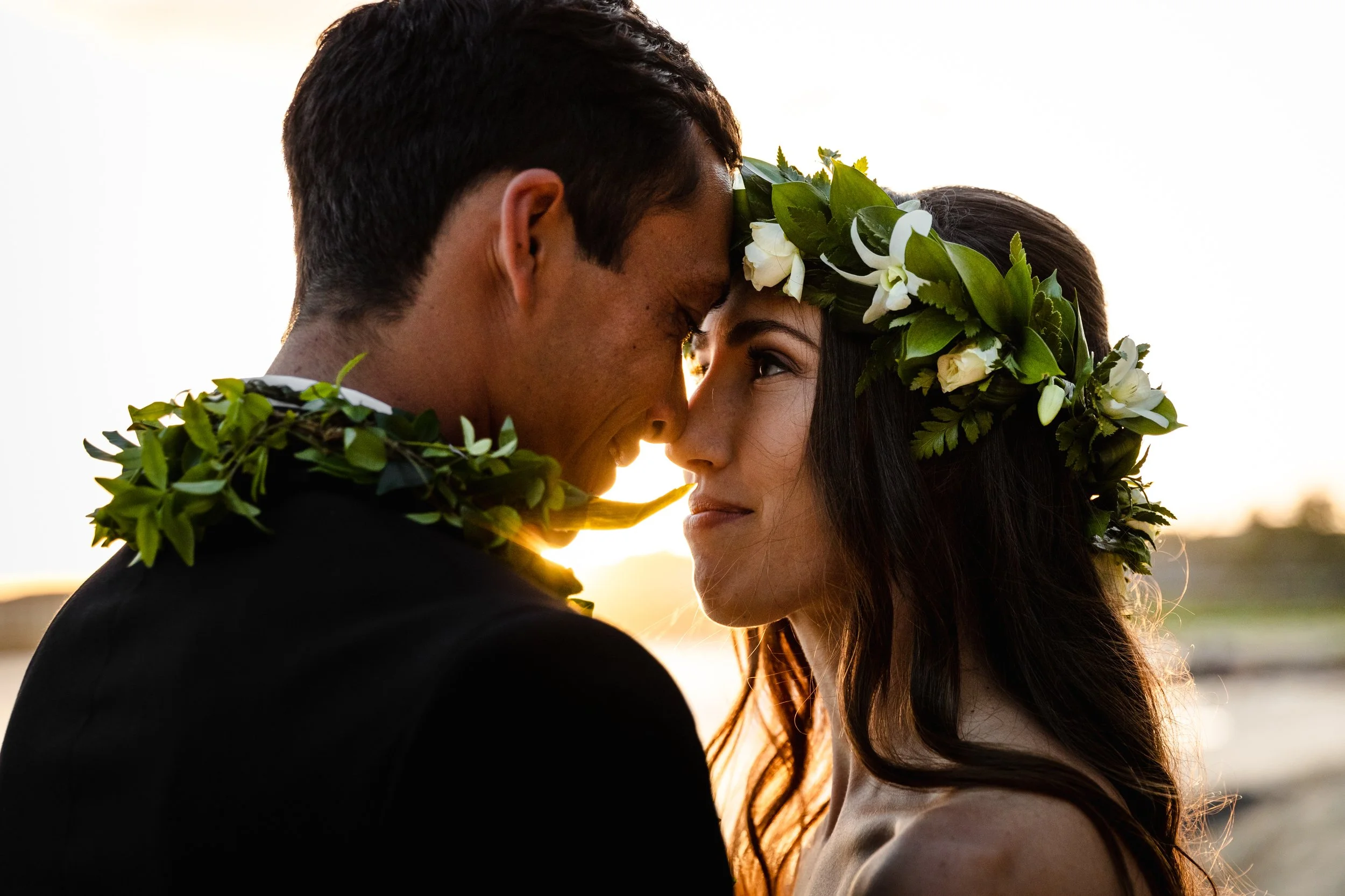 A man and woman are sharing a close moment, touching foreheads and smiling, with a floral crown on the woman's head, during sunsetPainting class on kauai instructed
