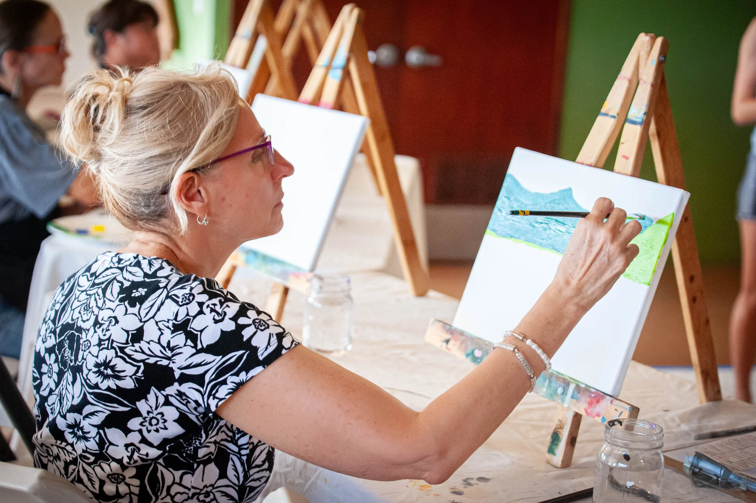 An older woman with gray hair tied up in a bun, wearing glasses and a black and white floral shirt, painting a landscape on a canvas. She is seated at a table with art supplies, in a room with other people also participating in a painting activity, with easels and canvases in the background