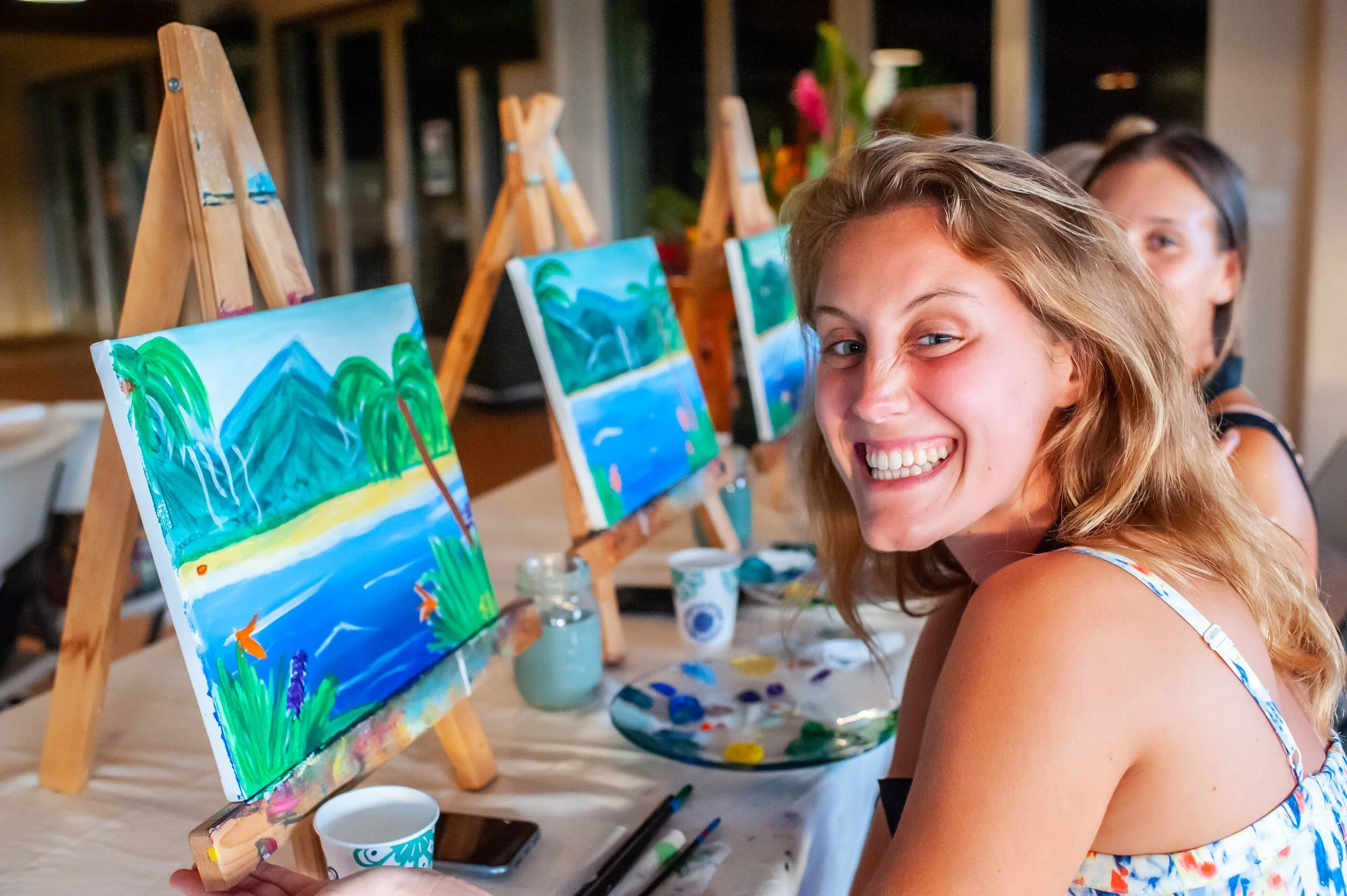 Smiling young woman with curly blonde hair painting landscape on canvas at an art class, with easels and paintings in the backgroundPainting class on kauai instructed