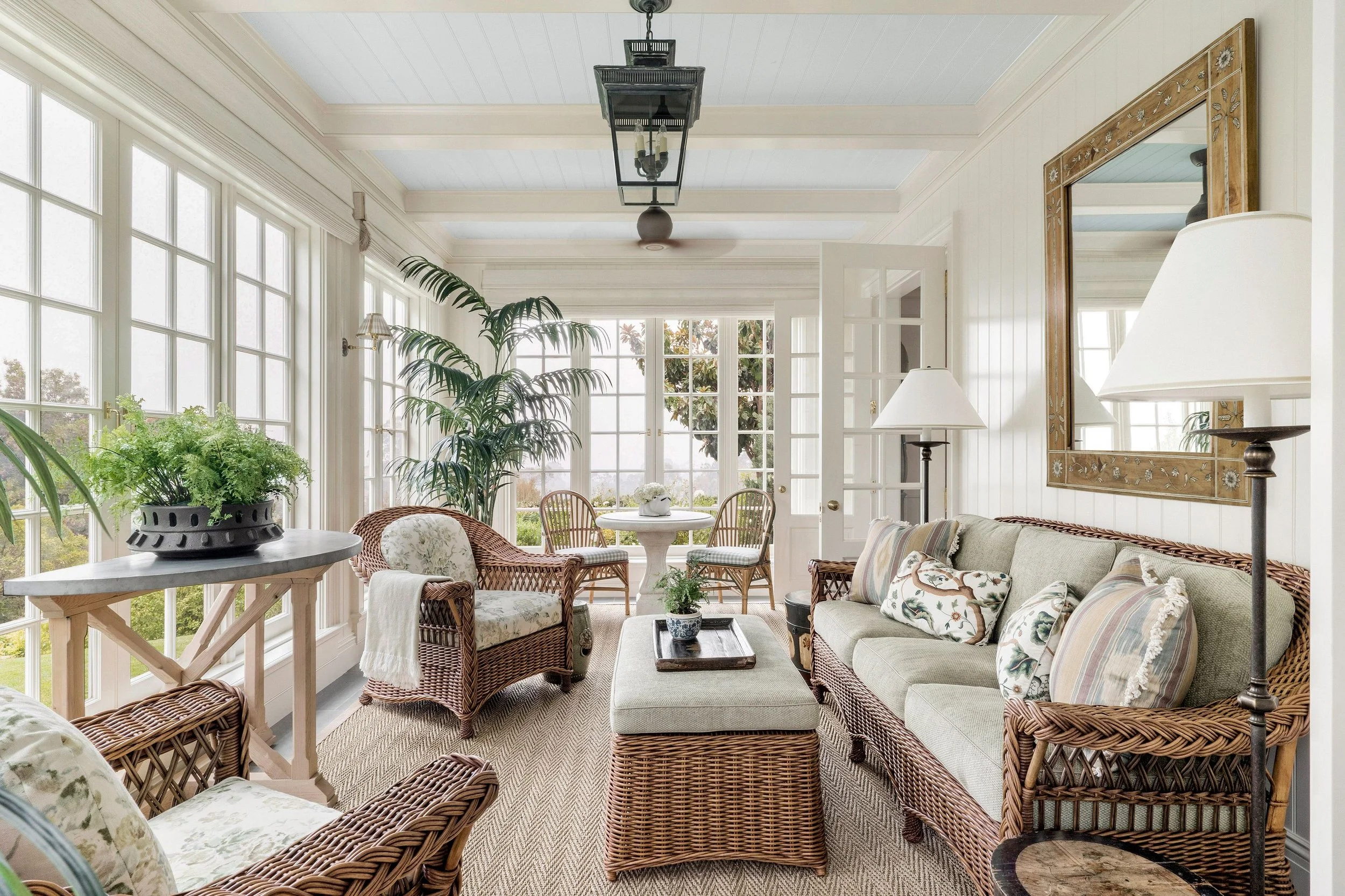 Sunlit enclosed porch with white walls, large windows, and ceiling. Features wicker seating with floral cushions, a white round table with a vase, potted plants, a large mirror, and lamps, creating a cozy, outdoor-inspired living space.