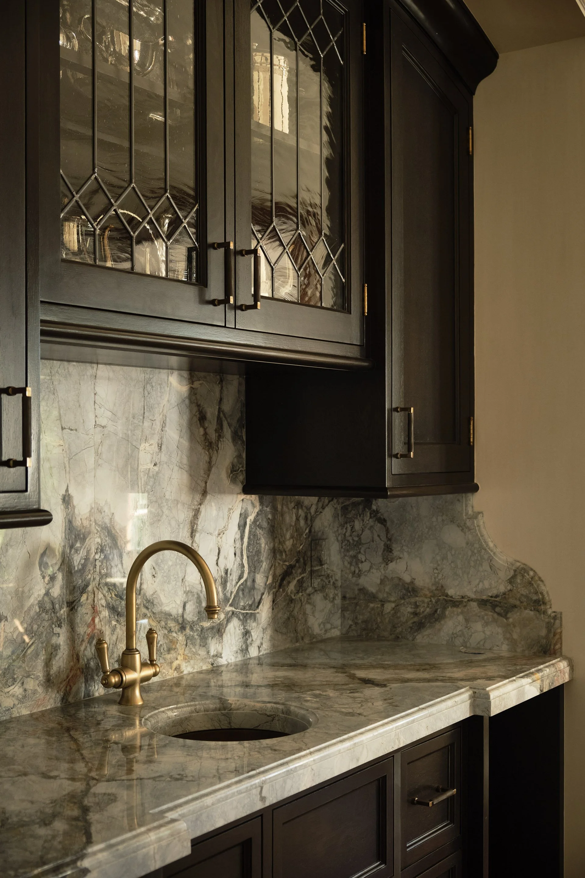 Kitchen detail with marble backsplash, brass faucet, and glass-front dark cabinetry