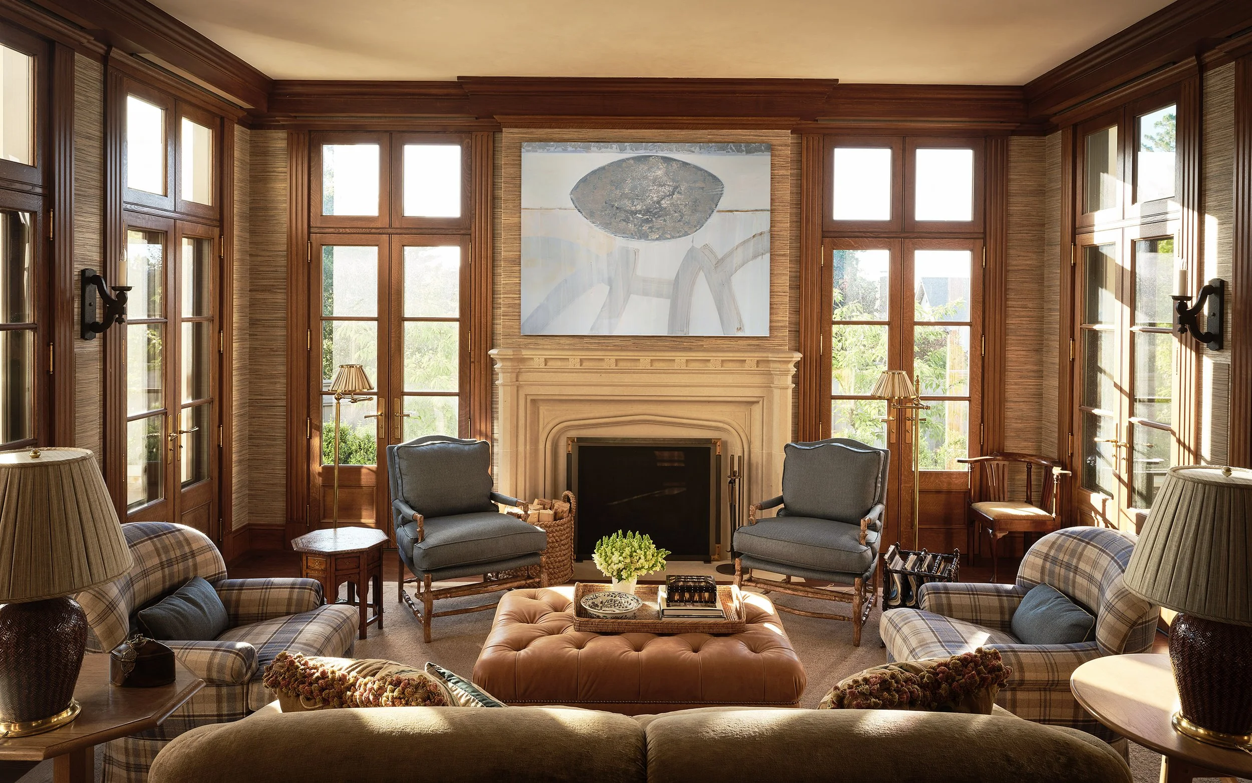 Living room with large windows, wood trim, a fireplace, artwork above the fireplace, armchairs, a plaid sofa, and various lamps and decor.