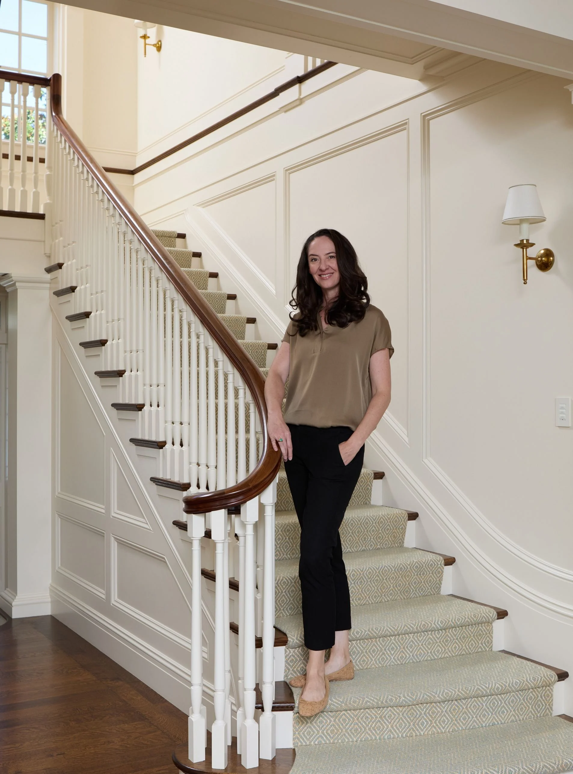 A woman stands on a staircase with white balusters, a wooden handrail, and beige patterned carpet. She is wearing a brown blouse and black pants, smiling, and indoors near a wall-mounted lamp.