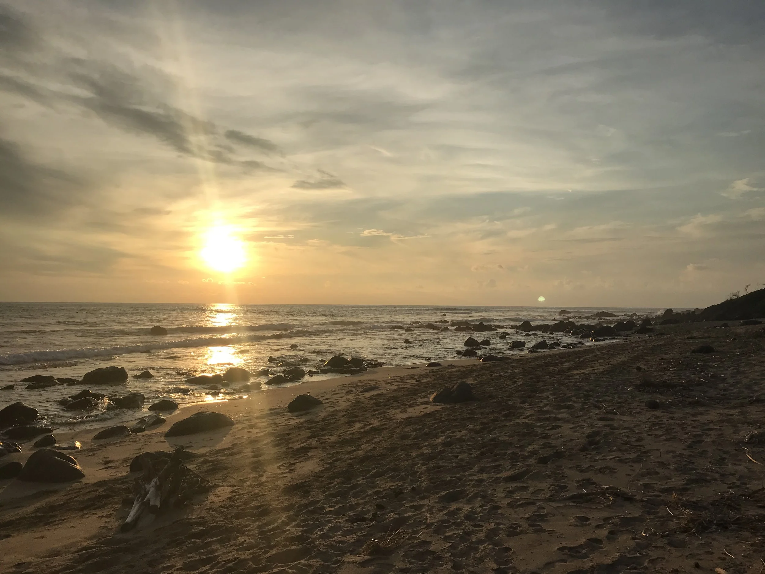 Sunset over the ocean with a sandy beach and scattered rocks in the foreground, partly cloudy sky with a rainbow.