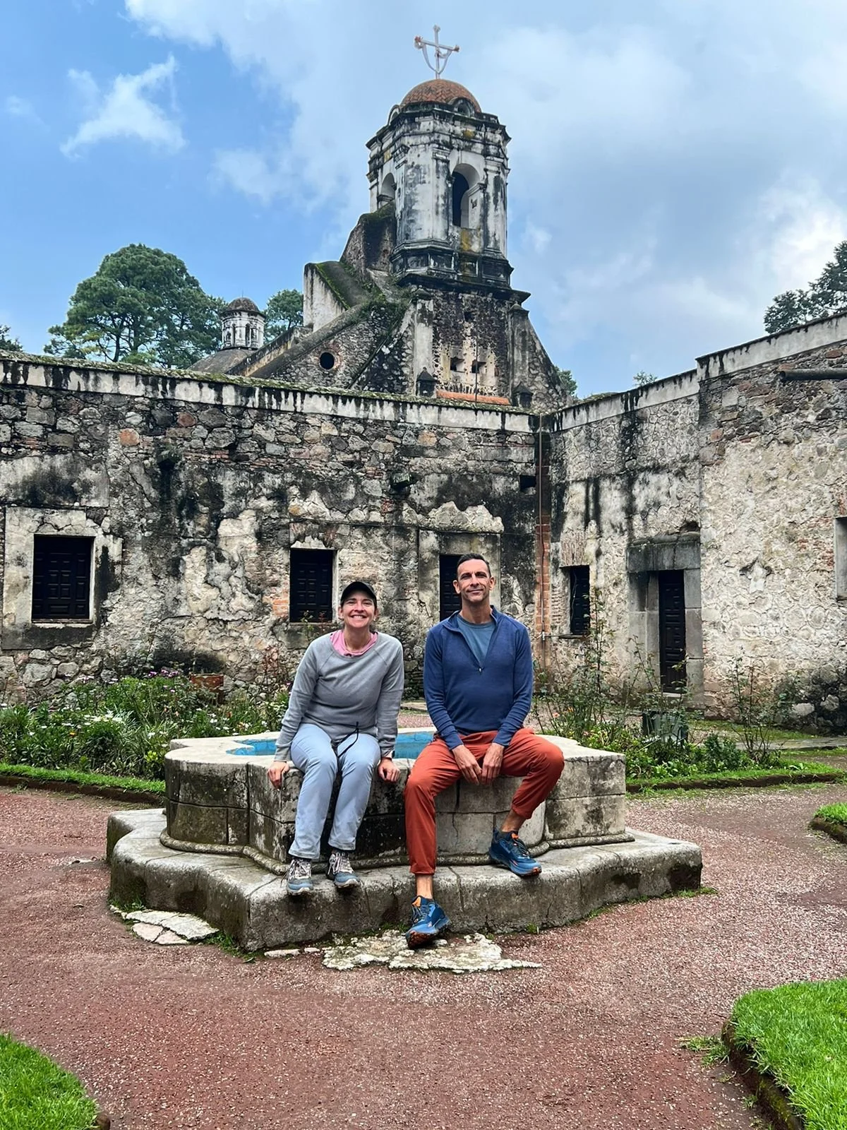 Two people sitting on a stone bench in front of an old stone building with a tower and a cross on top, surrounded by greenery and a cloudy sky.
