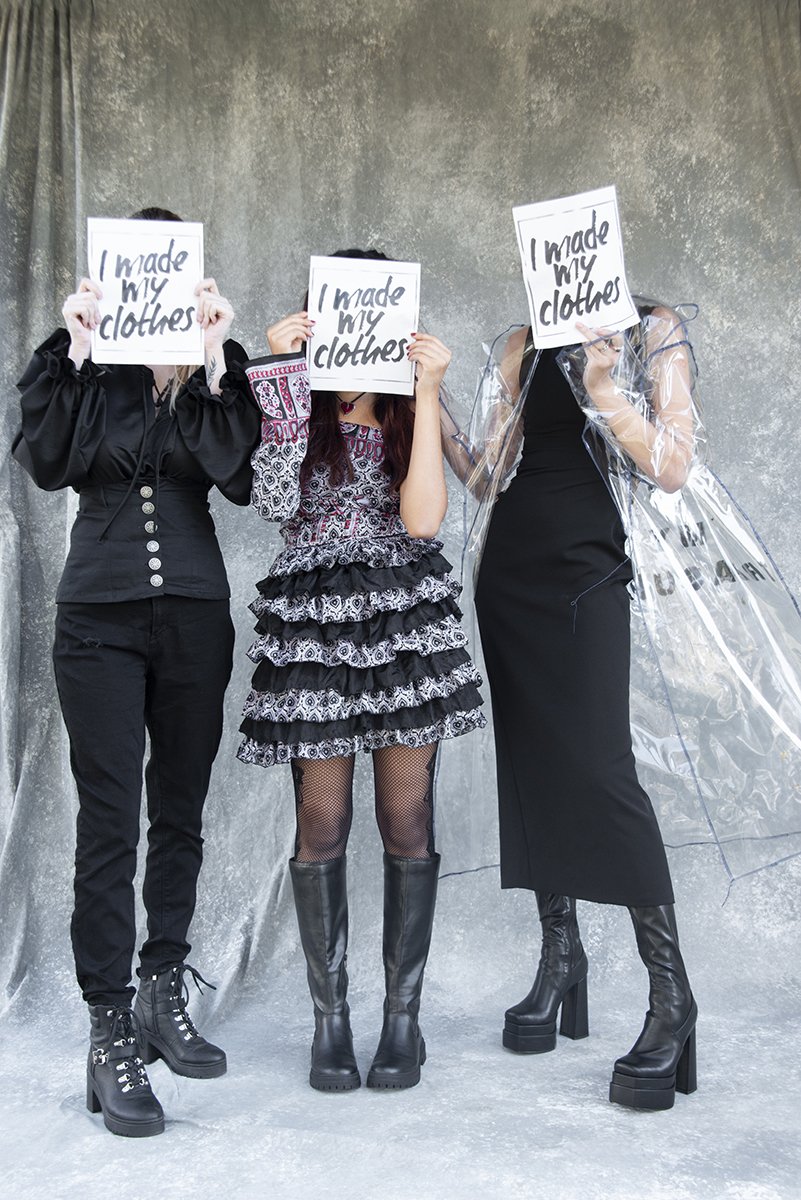 Three women standing in front of a gray textured backdrop, holding signs that say 'I made my clothes' covering their faces. The woman on the left wears black pants, a black jacket, and black boots. The woman in the middle wears a patterned ruffled dress, fishnet stockings, and tall black boots. The woman on the right wears a black dress, clear plastic cape, and black platform boots.